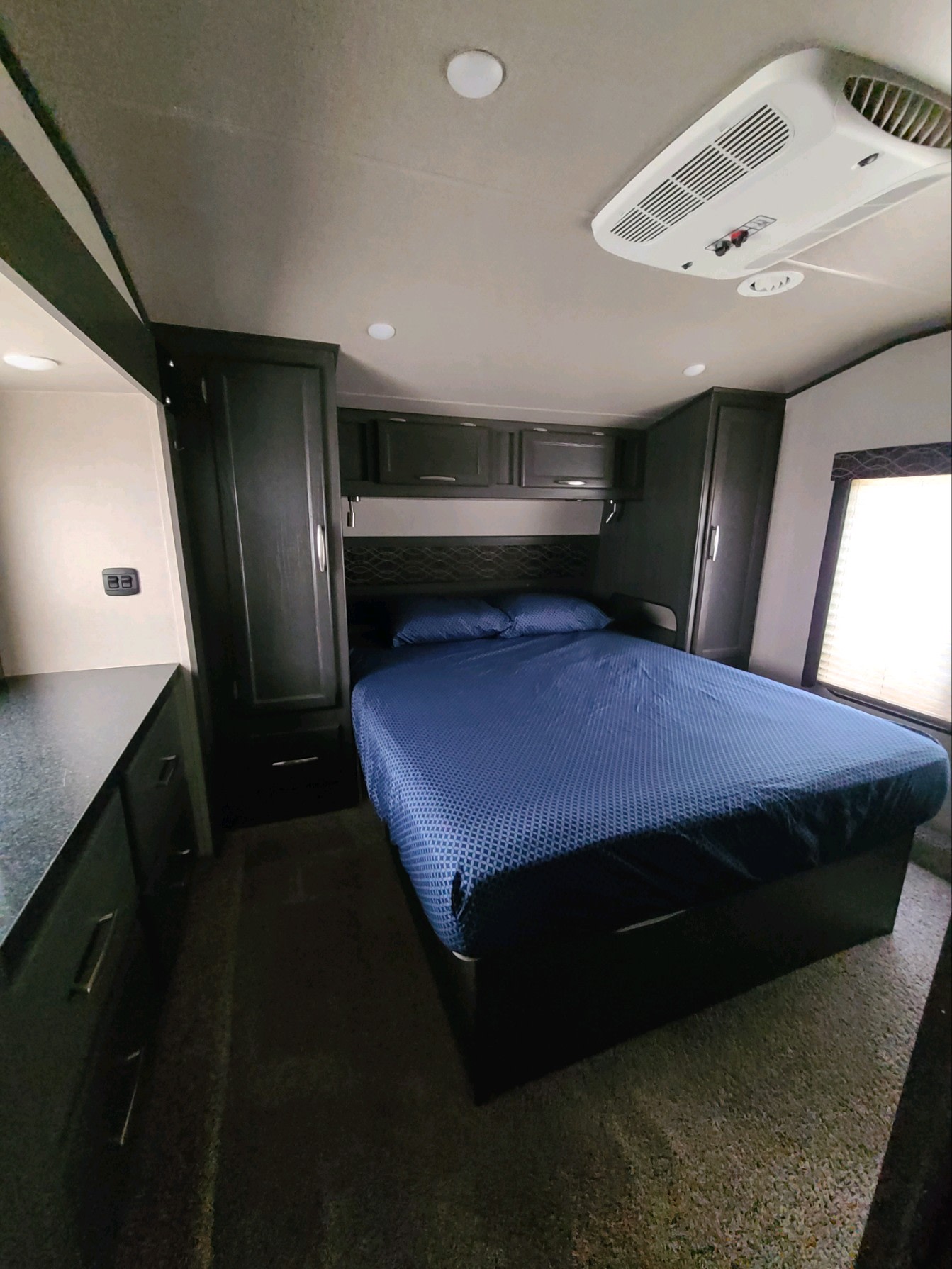 Comfortable RV rental bedroom: blue bed, dark cabinetry, AC, & window. Ideal for road trips & adventure.. Genesis Supreme Toy Hauler 2019