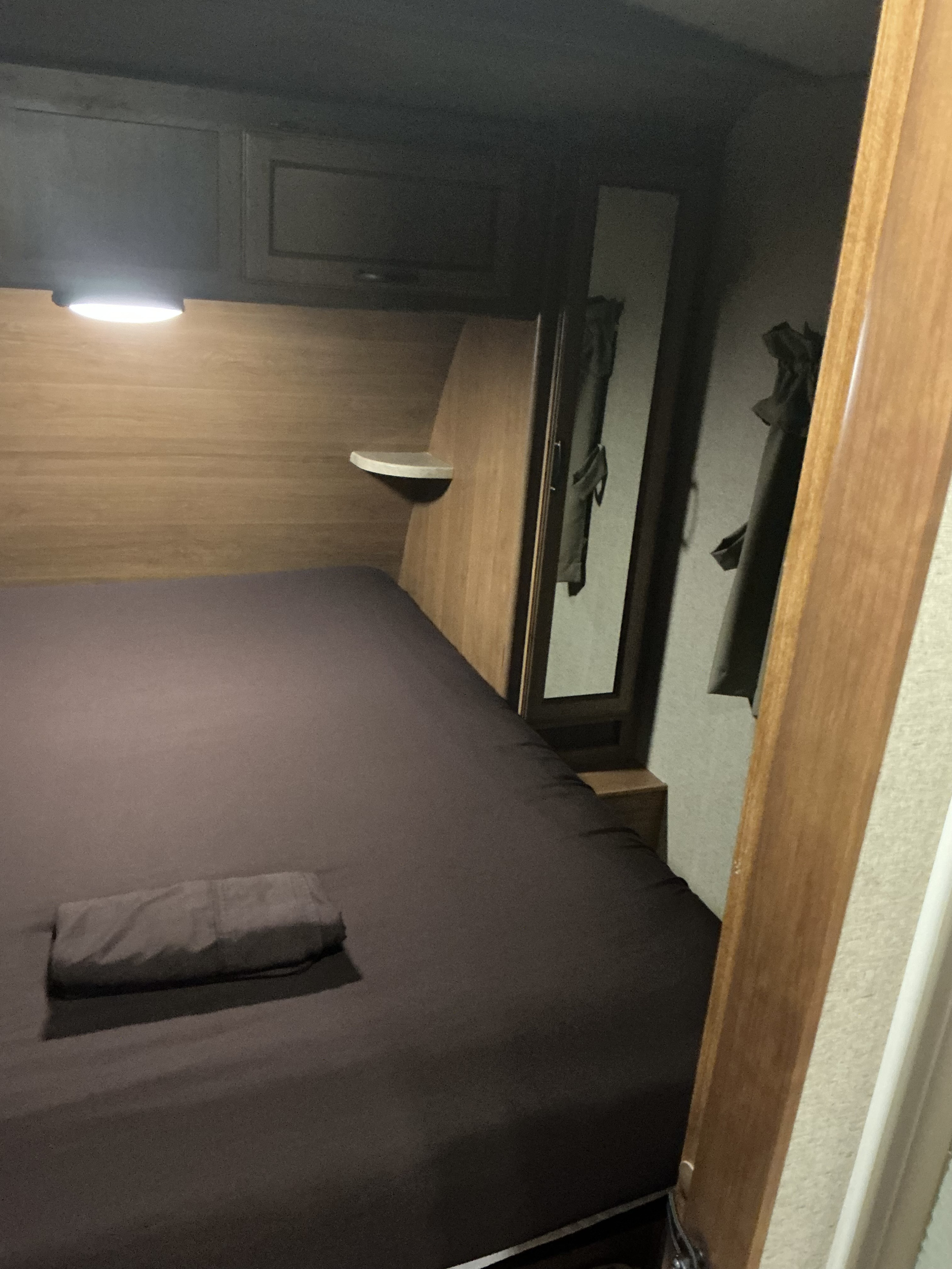 An RV rental bedroom shows a dark brown bed with folded linen, bright overhead light, and a small side shelf. Compact space.. Crossroads RV Sunset Trail Super Lite 2017