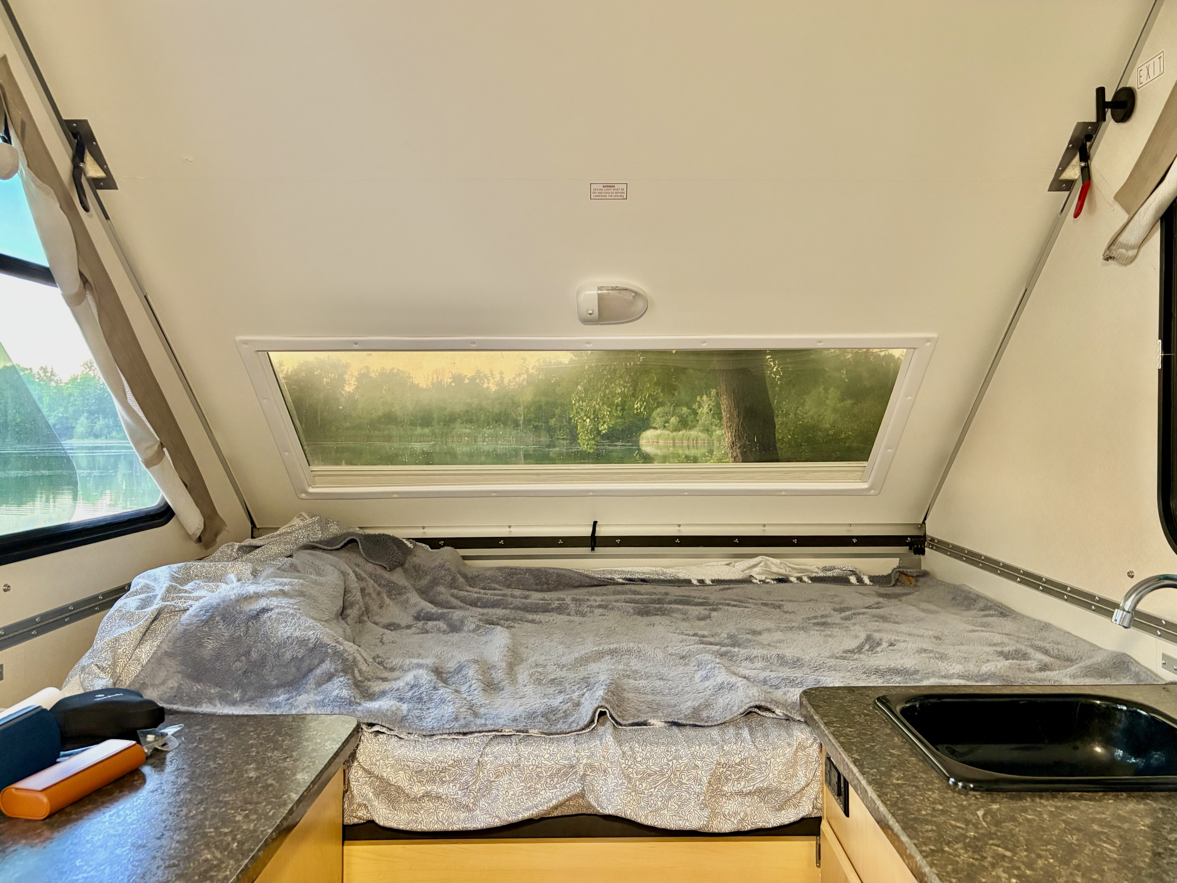 Cozy RV rental interior with a comfortable bed, compact kitchen area, and a scenic window view of a lake and lush trees.. A Liner A Liner Popup Trailer 2021