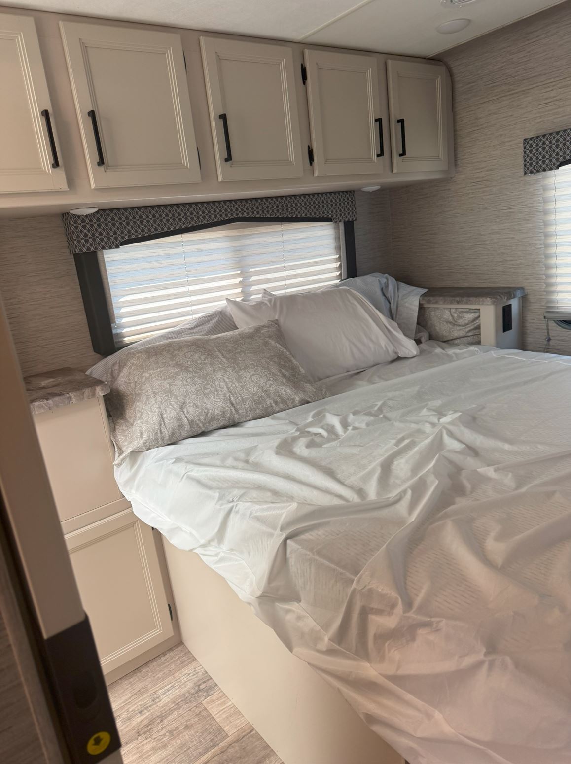 Cozy RV rental bedroom with a bed, white linens, overhead cabinets, and a window with horizontal blinds.. Thor Chateau 2023