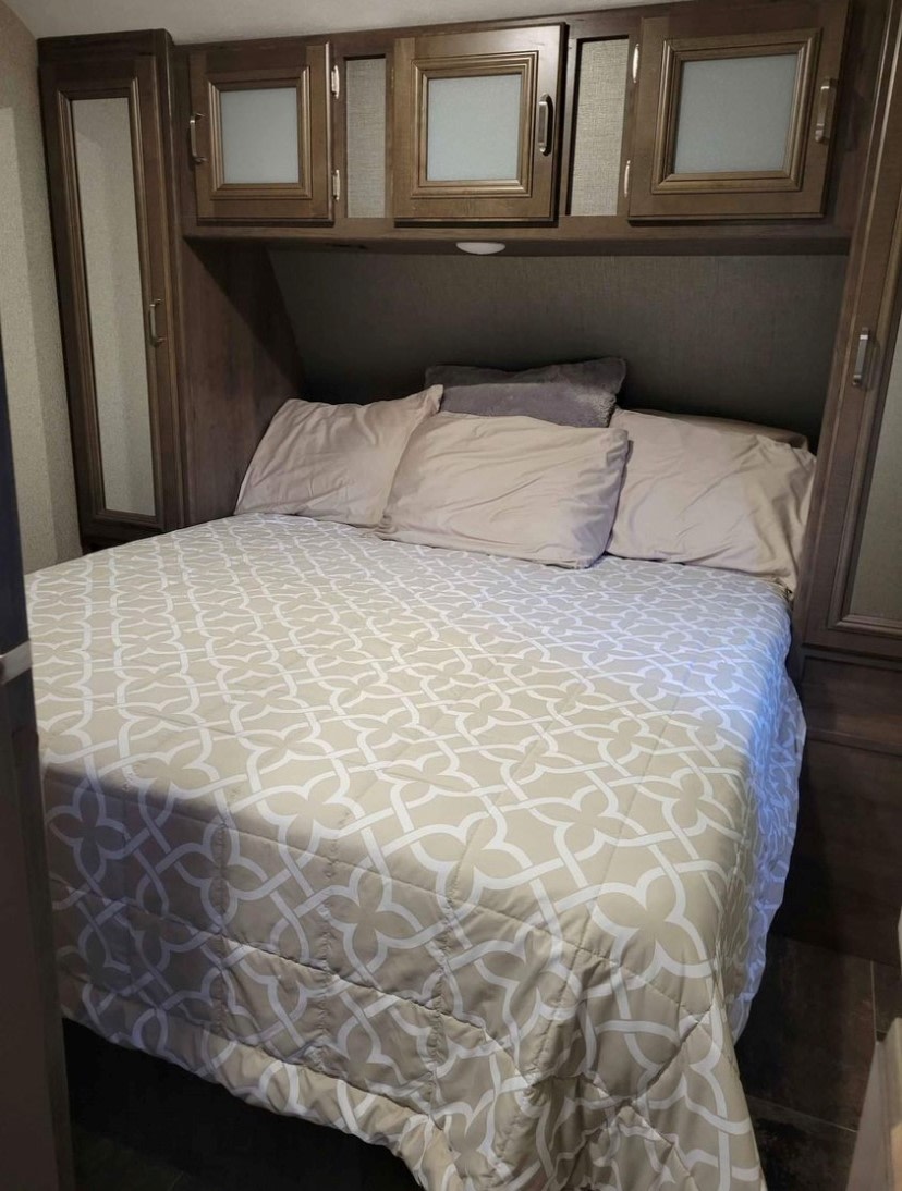 RV rental bedroom: a bed with patterned bedding, pillows, plus overhead and side wooden cabinets.. Keystone RV Passport Grand Touring 2019