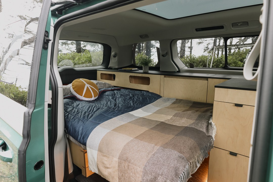 Inside a comfy RV rental: bed with peace sign pillow, wood cabinets, and nature views through the windows.. Volkswagen Other 2025