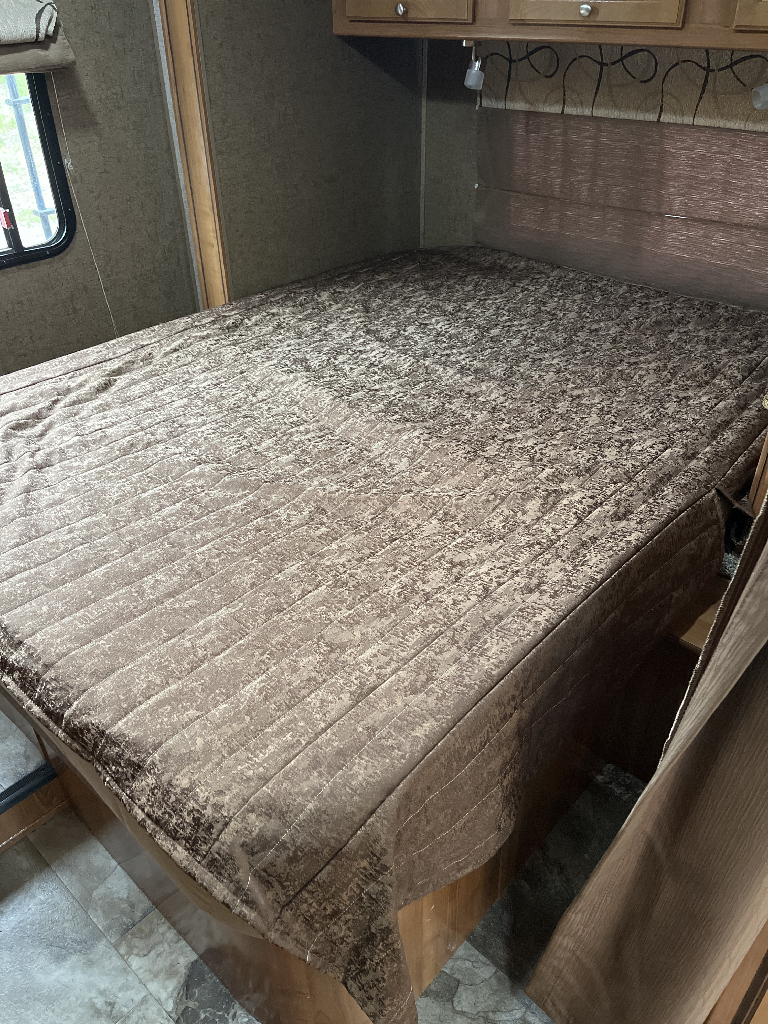 RV rental bedroom with patterned bed covering, overhead cabinets, and window, providing a cozy interior.. Forest River Coachmen Leprechaun 2018