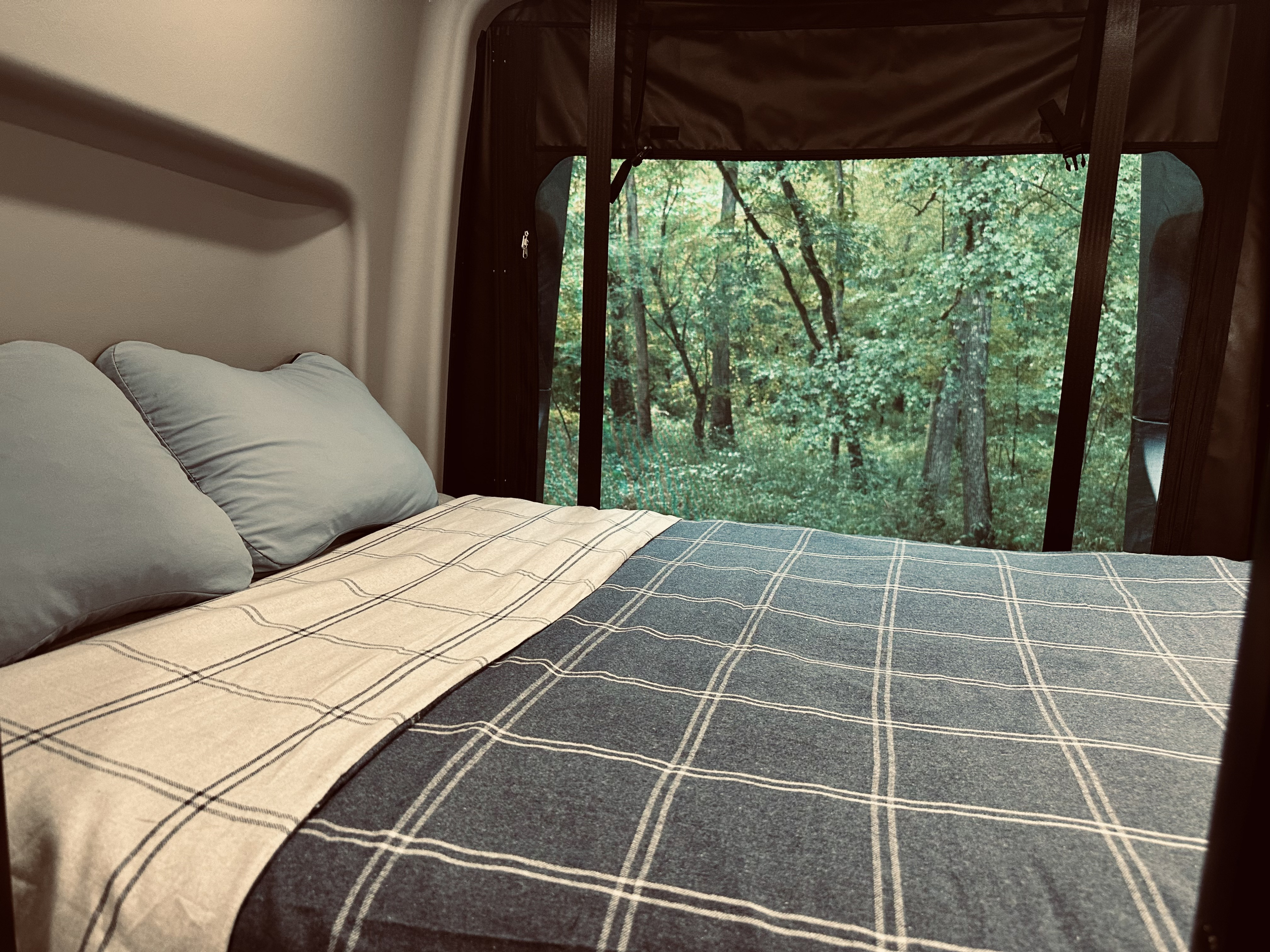 Comfortable RV rental bed with a serene forest view from the large window, perfect for camping and nature lovers.. Winnebago Revel 2025