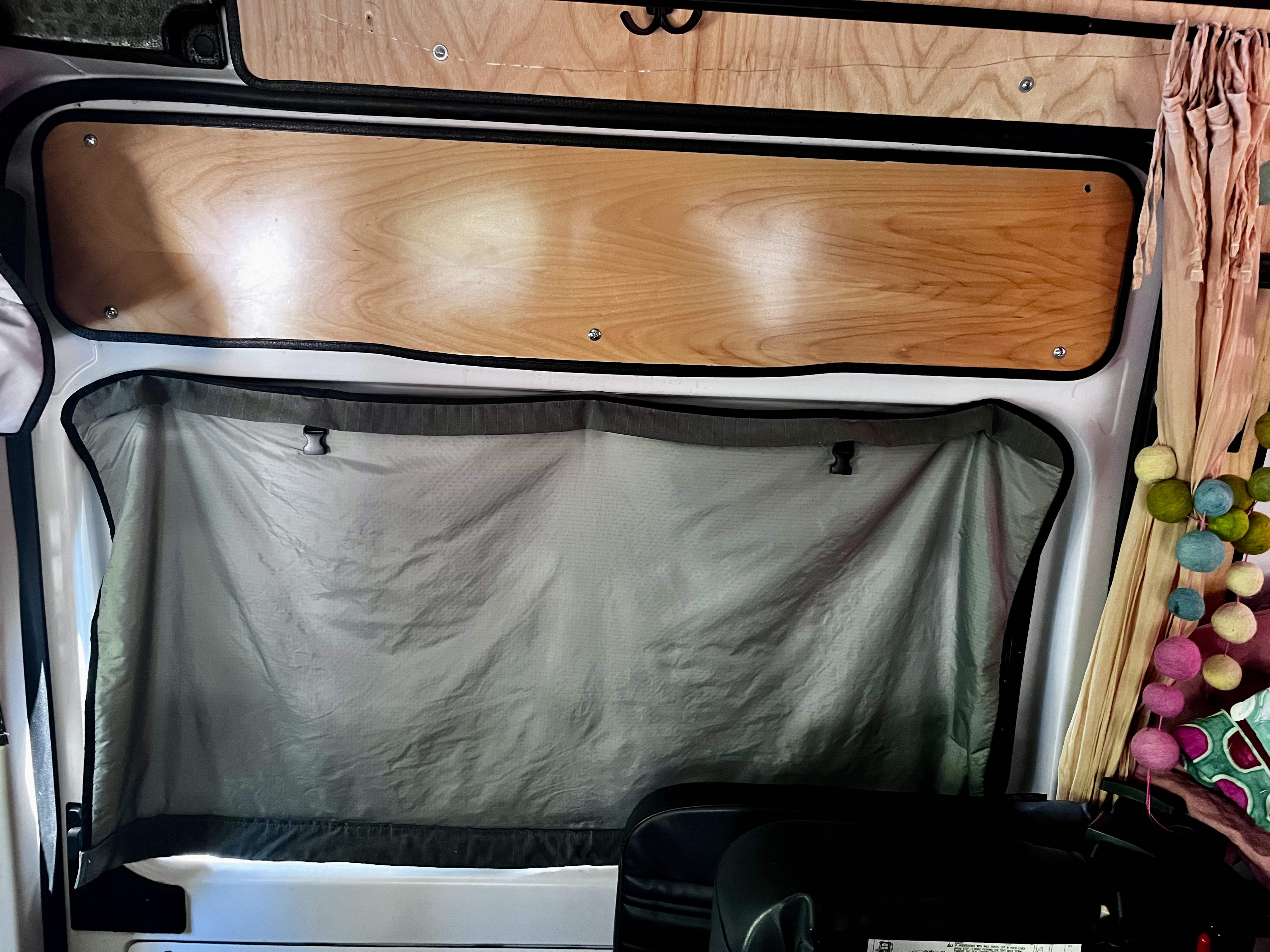 Interior of an RV rental, showing a window with a grey privacy shade, wooden paneling, and decorative felt balls for a cozy camper aesthetic.. Dodge Ram Promaster 2500 2021