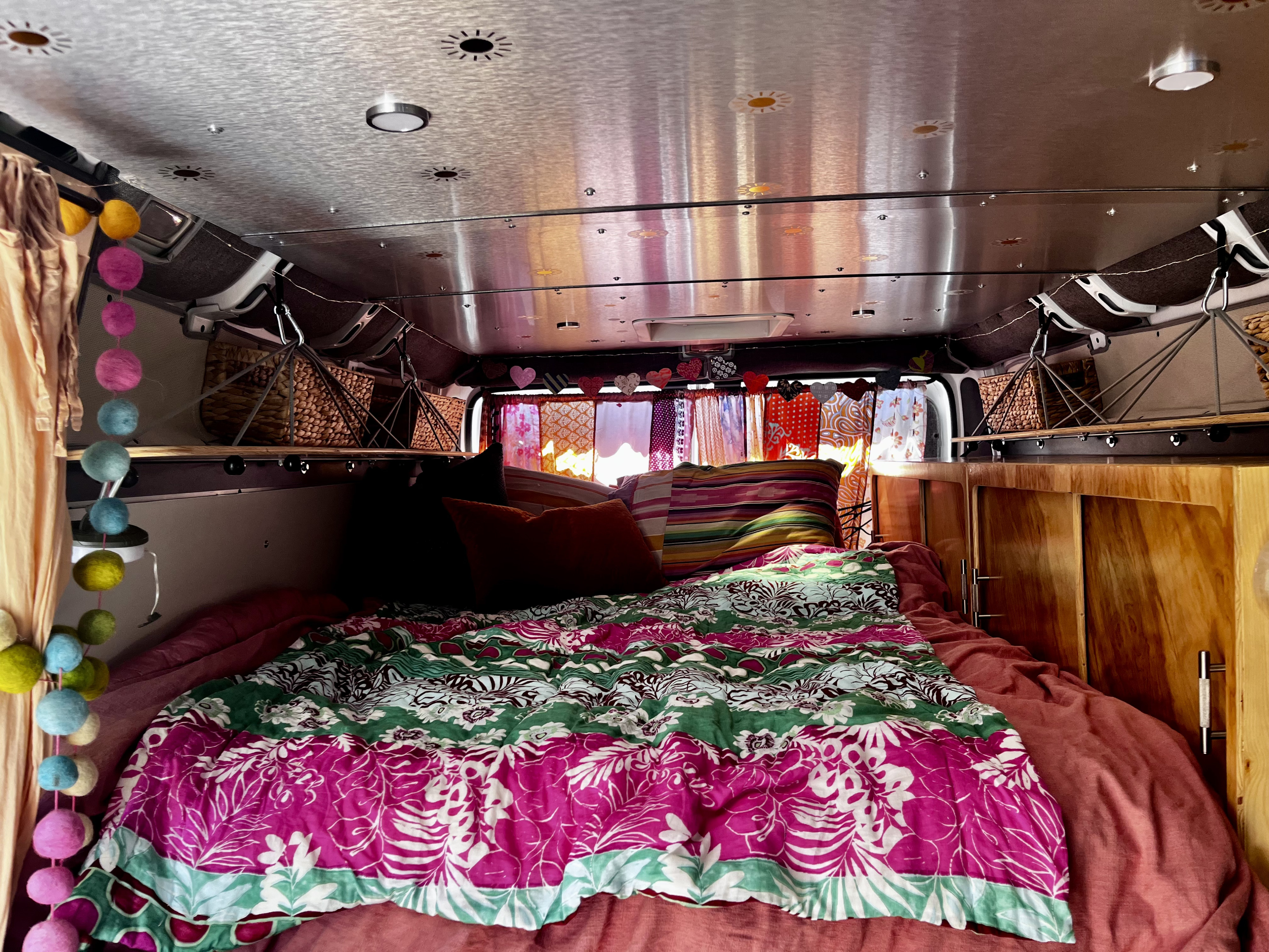Inside a vibrant RV rental: cozy bed with pink/green quilt, wooden cabinets, and string lights create a warm, inviting space.. Dodge Ram Promaster 2500 2021