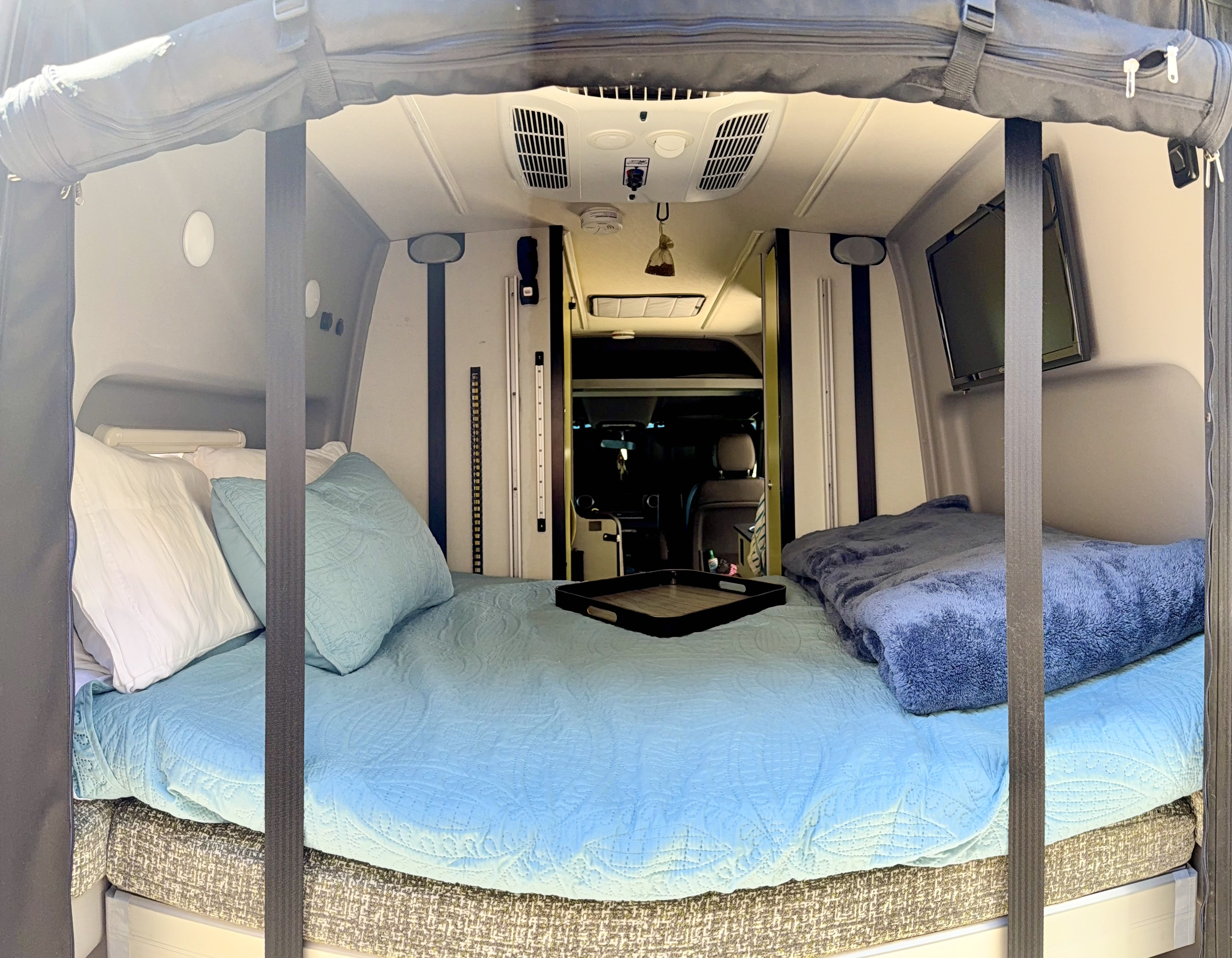 Inside a cozy rv rental: A comfortable bed with light blue bedding, pillows, a tray, and a TV awaits travelers.. Winnebago Revel 2020