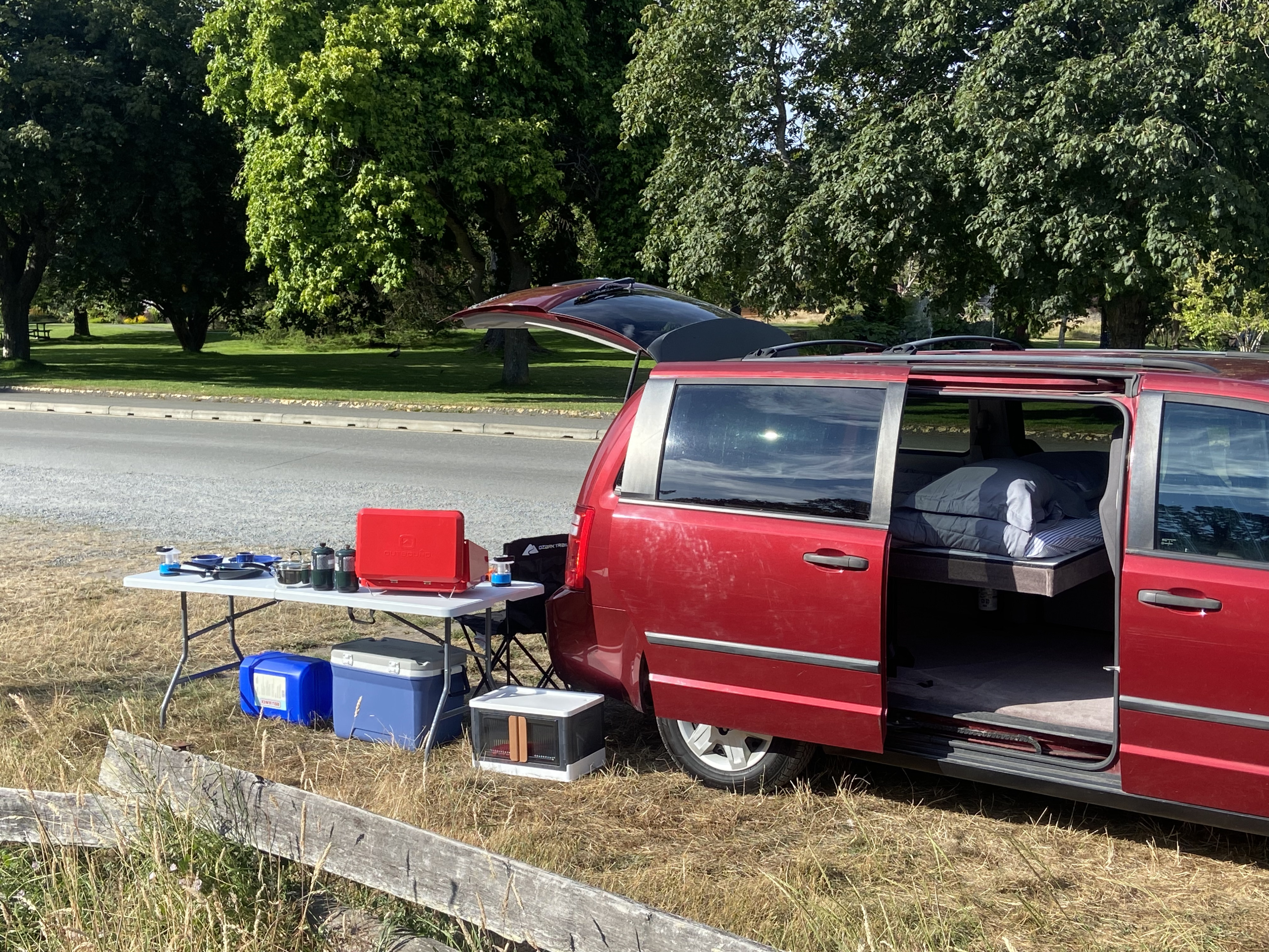 This red minivan camper conversion, like an RV rental, is set up in a park with a cooking table and a bed.. Dodge Grand Caravan 2010