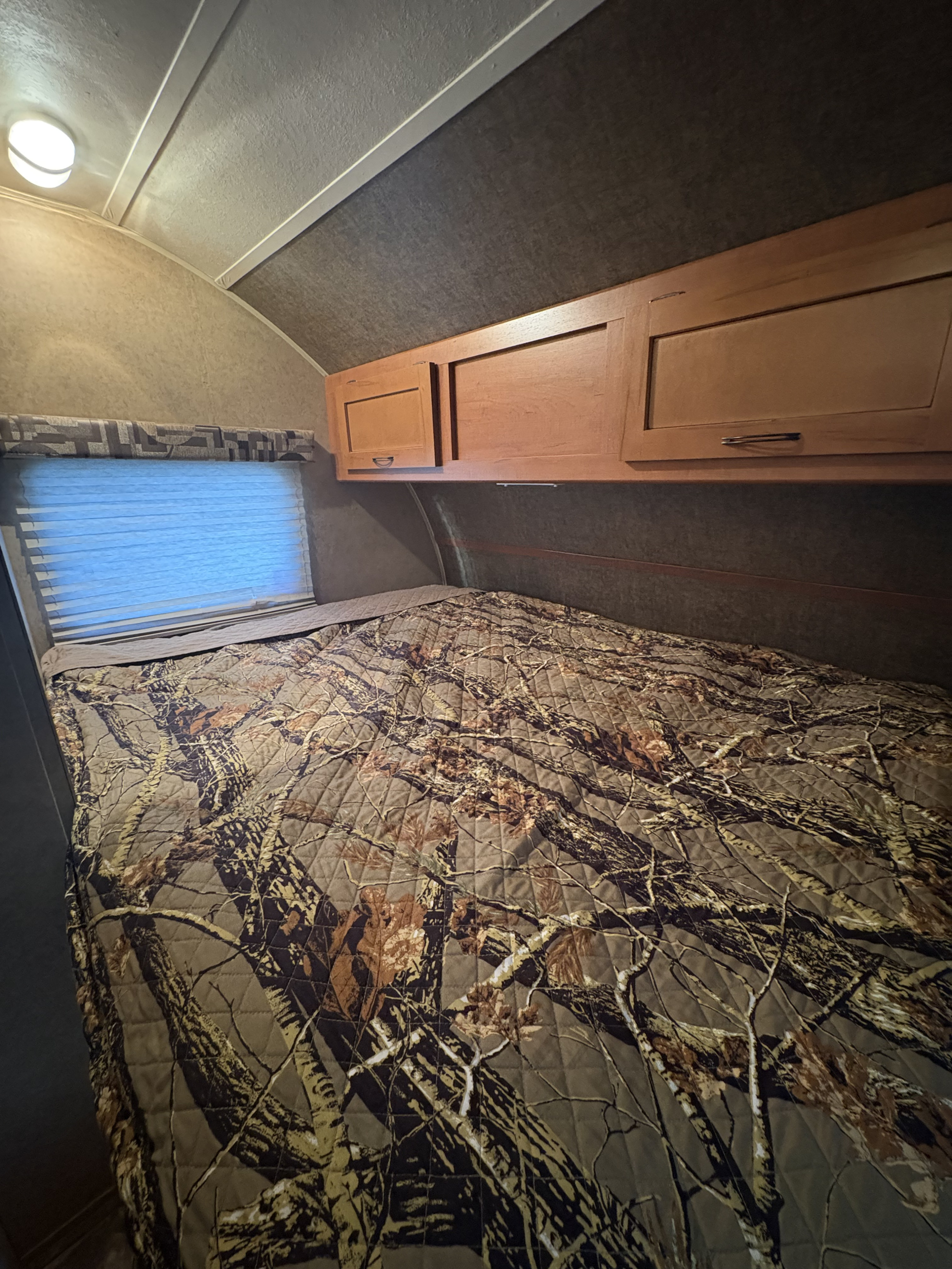 Cozy RV rental bedroom with a bed featuring a detailed camouflage pattern, window, and wooden overhead cabinets.. Forest River Cherokee Grey Wolf Toy Hauler 2014