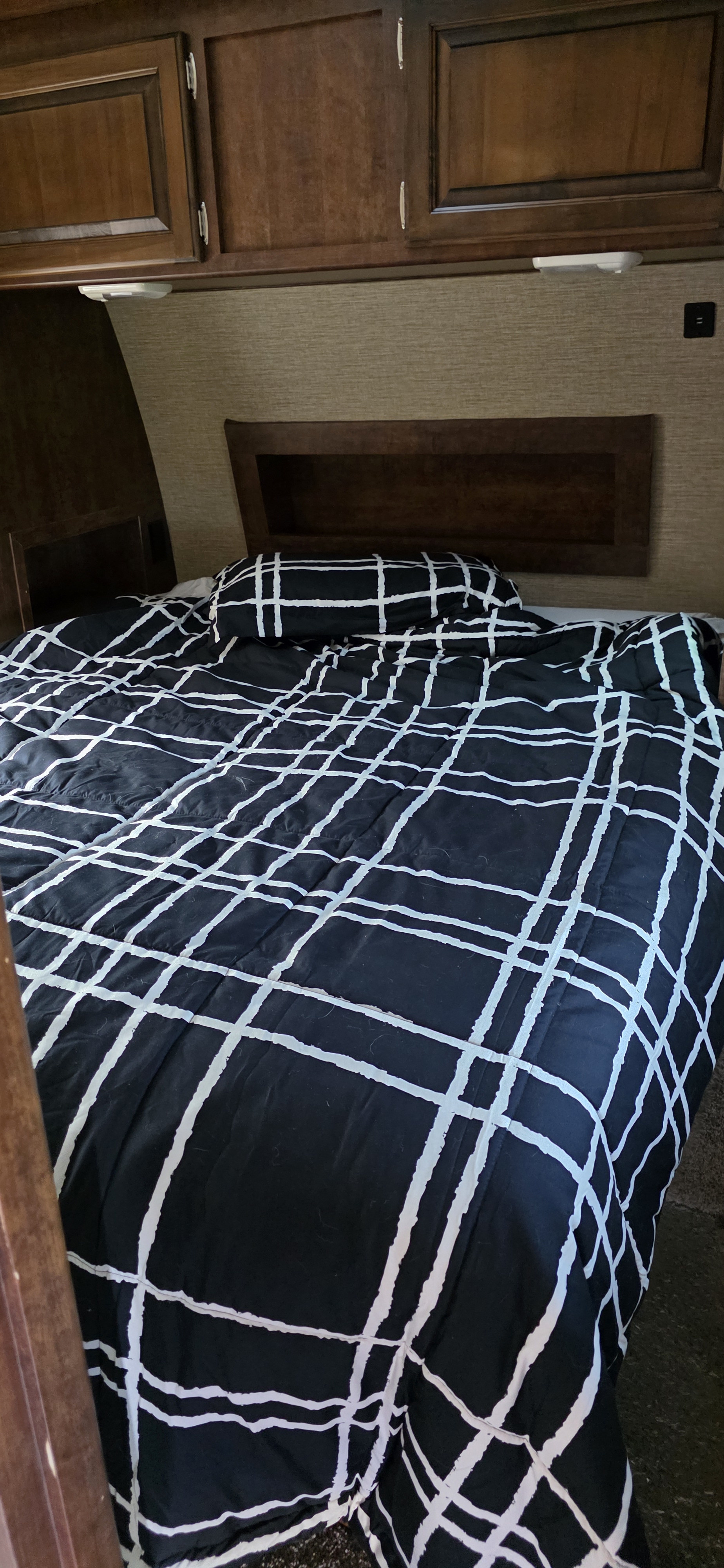 Cozy RV rental bedroom features a bed with a black and white grid comforter, wooden cabinets, and headboard.. Keystone RV Outback Super-Lite 2017