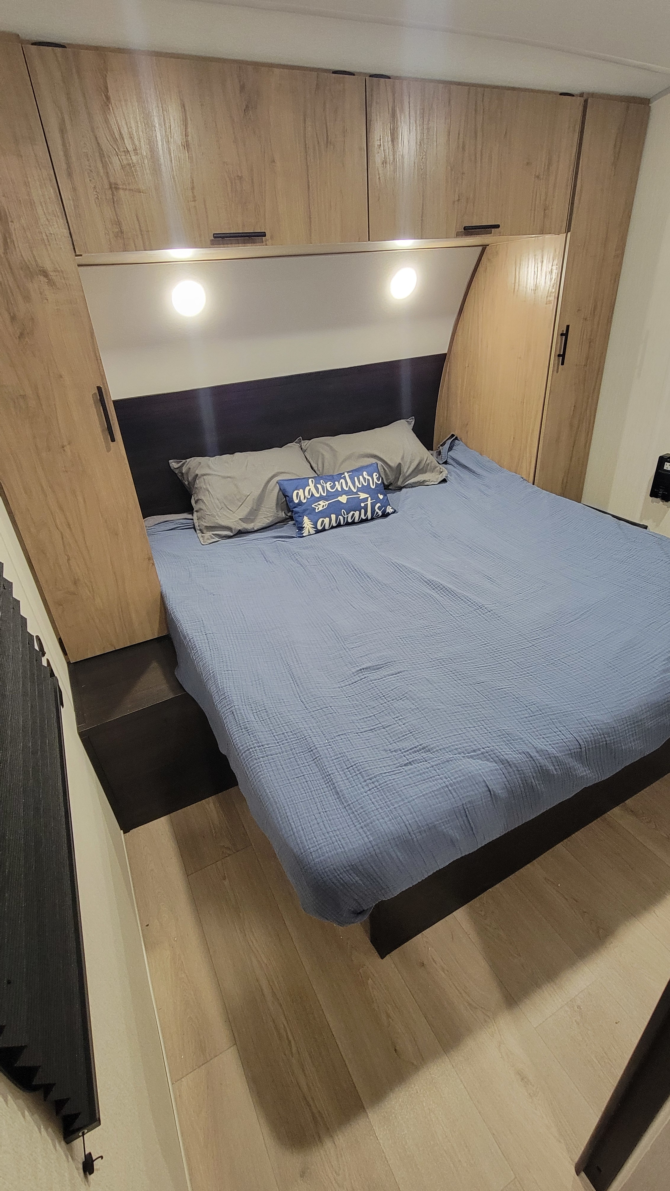 View of a modern RV rental bedroom showcasing light wood cabinets, reading lights, blue bedding, and an 'adventure awaits' pillow.. Cherokee Wolf Den 294GEBG 2025