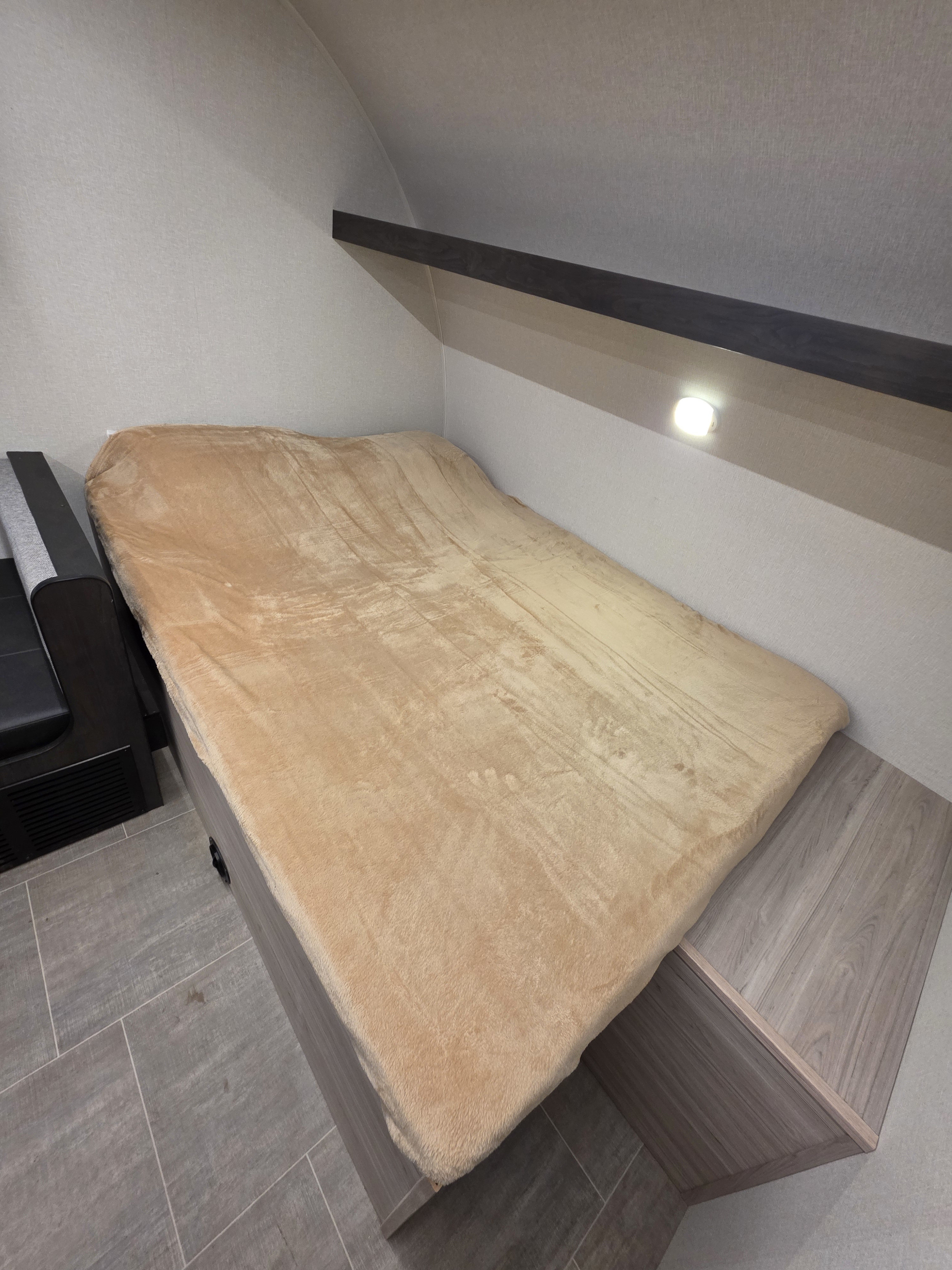 Inviting RV rental sleeping nook featuring a soft tan blanket, overhead shelf, and built-in light. Compact and functional.. Heartland Mallard Sport 2025