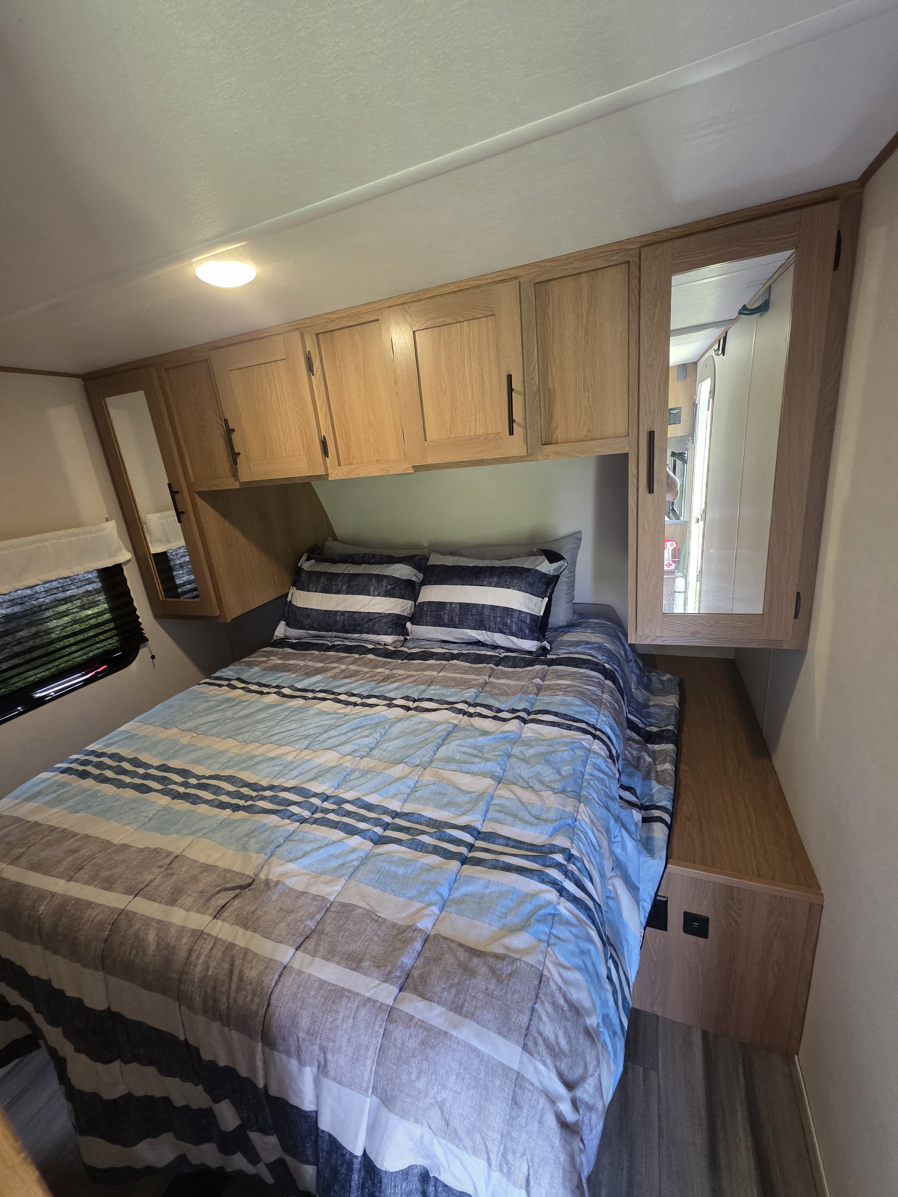 RV rental bedroom with a bed, blue striped bedding, overhead wood cabinets, and a mirrored closet door.. Avenger Avenger Trailer 2022