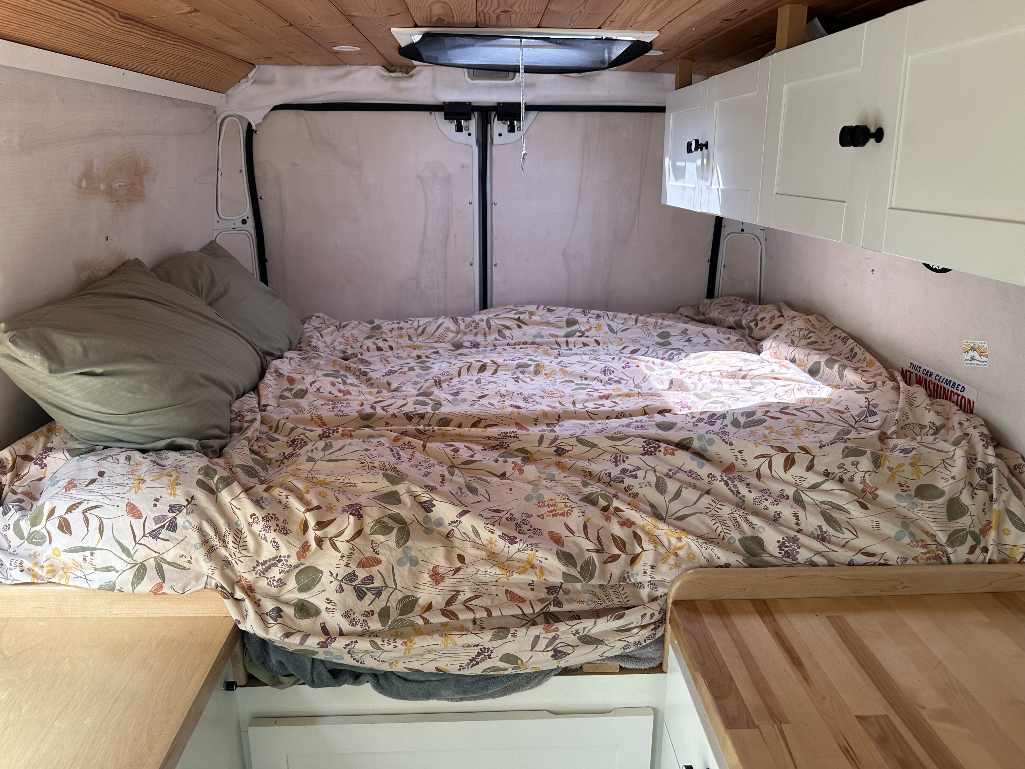 Cozy RV rental: Van bed with floral duvet, wood walls, and overhead storage cabinets for comfortable travel.. Dodge B Van 2018