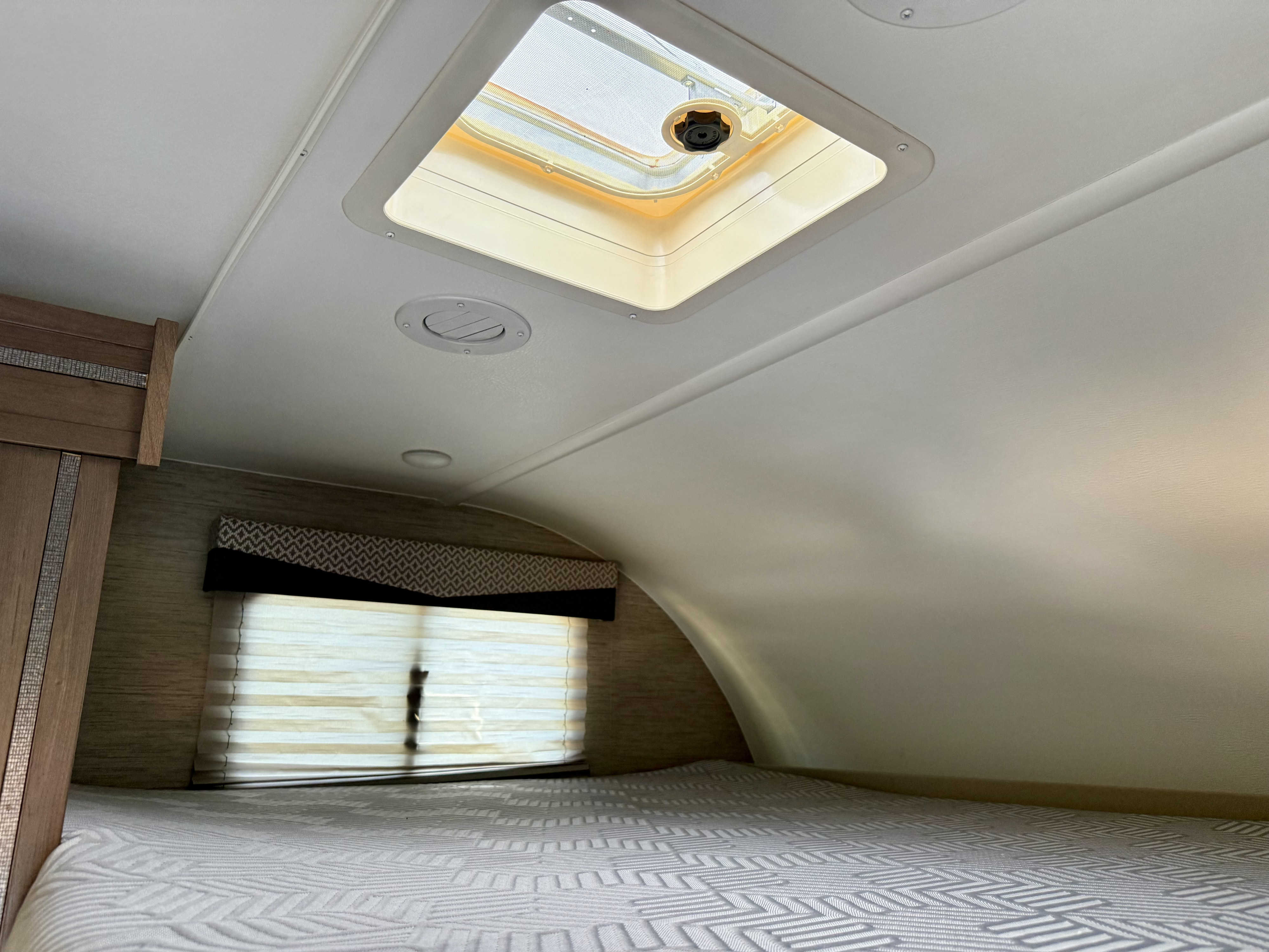 An RV rental's sleeping area, featuring a cozy bed, window with blinds, overhead vent, and bright skylight.. Thor Four Winds 2022