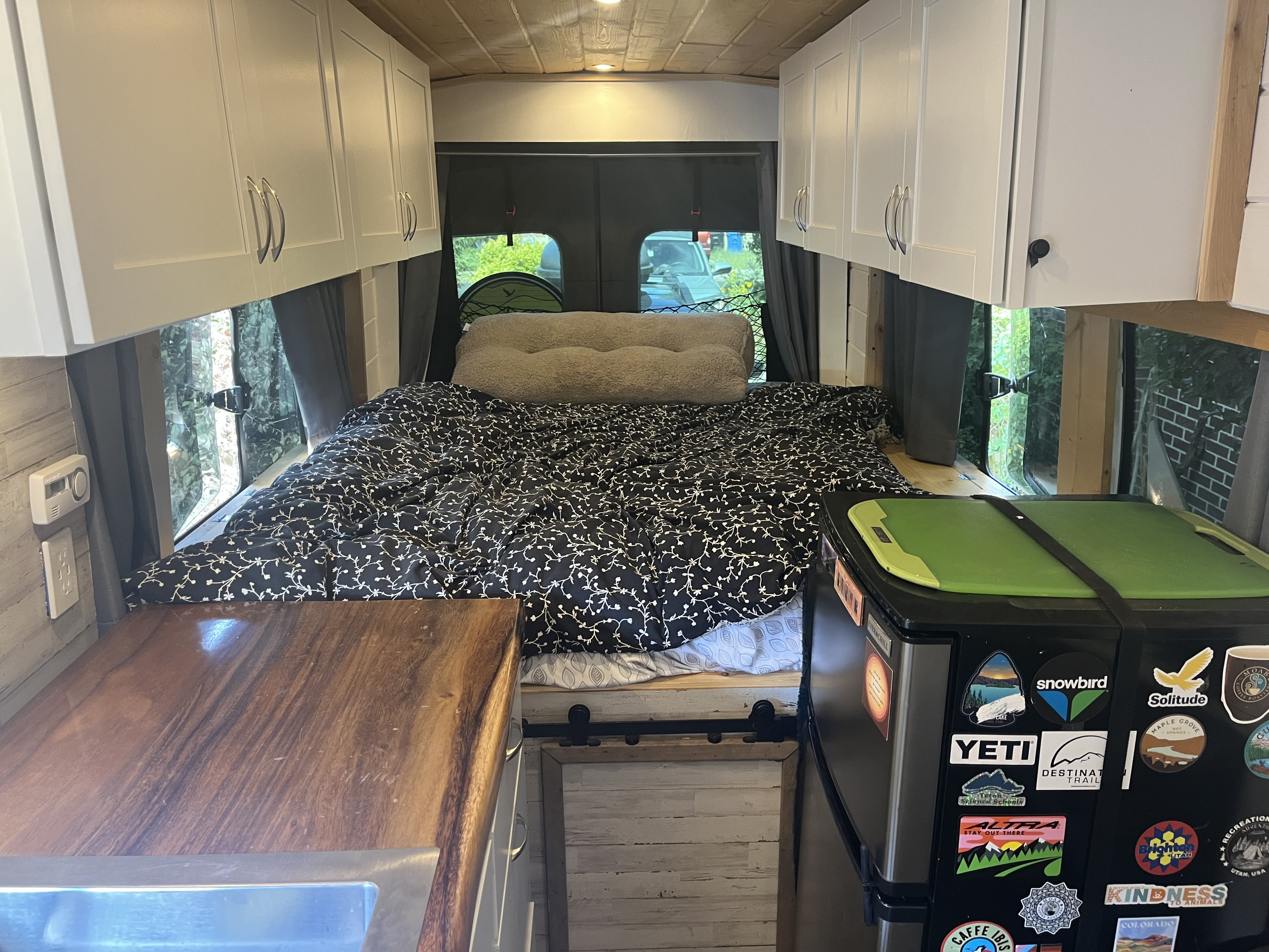 Cozy RV rental interior featuring a comfortable bed, integrated kitchen counter, overhead cabinets, and a fridge, perfect for mobile living.. Ford Transit 2016
