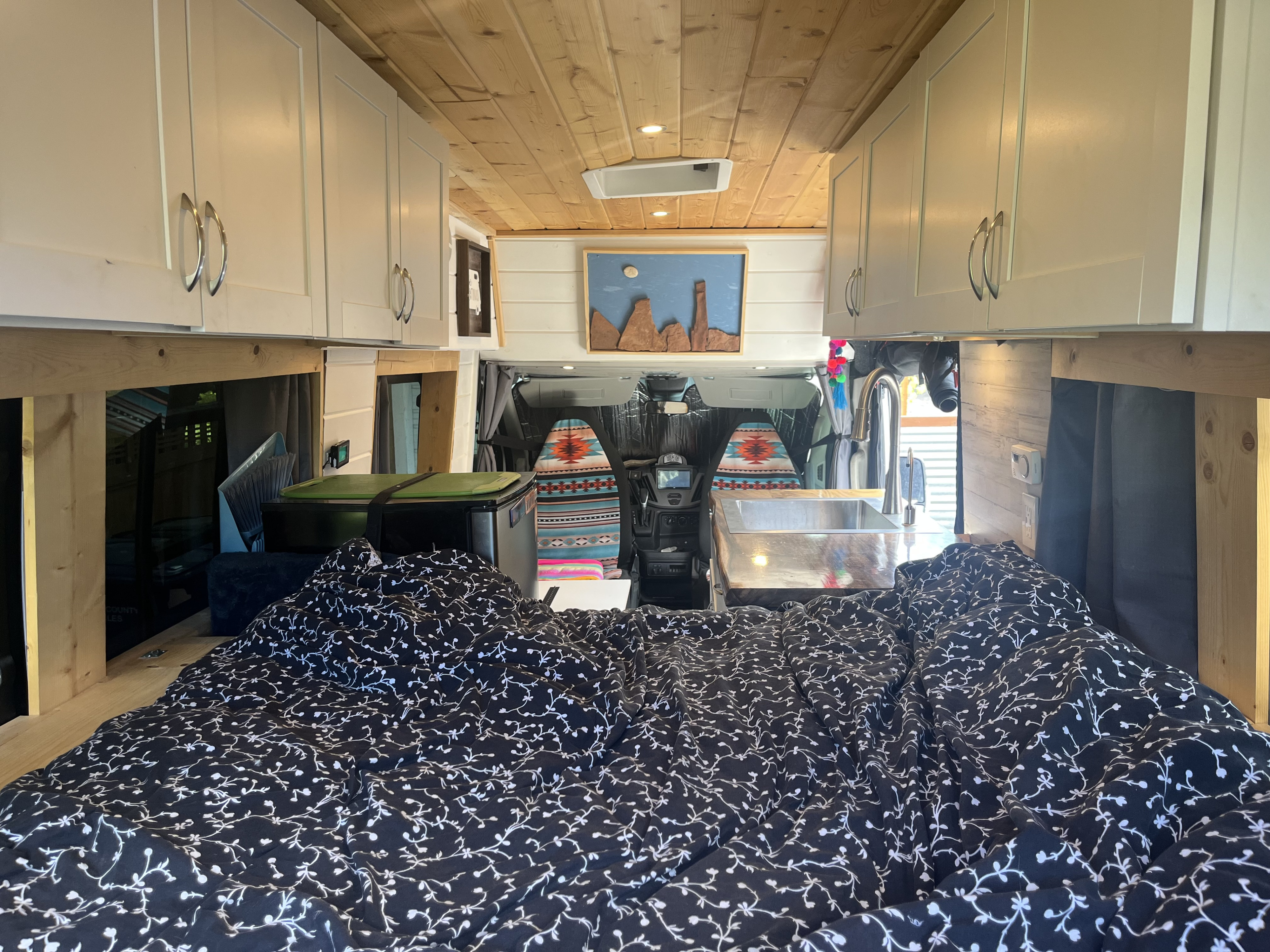 View of a camper van's interior from the bed towards the front, showing a kitchen sink, fridge, and overhead cabinets. Ideal for an RV rental.. Ford Transit 2016