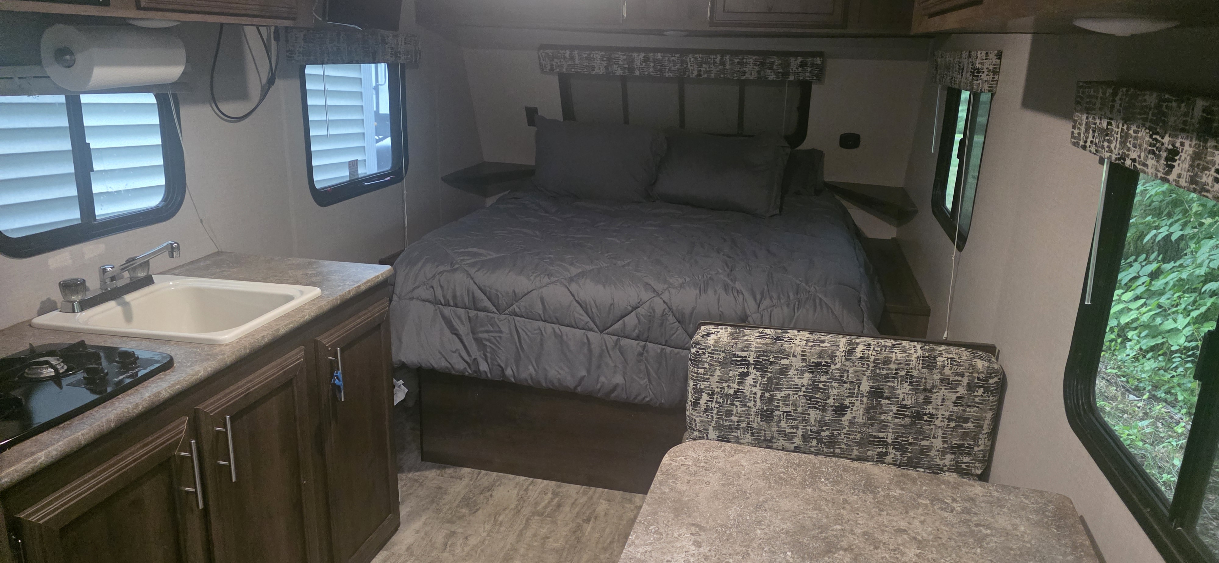 RV rental interior featuring a bed, kitchen area with sink/stove, and dinette. Windows show nature and nearby structures.. KZ Sportsman Classic 180 QB 2018