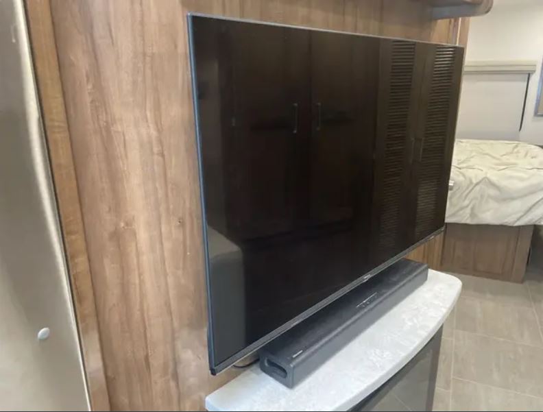 Modern RV rental interior with a large flat-screen TV and soundbar. Wood-paneled wall and partial bedroom view.. Forest River Coachmen Encore 2021
