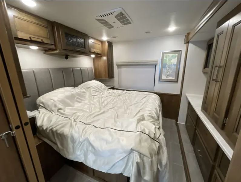 Inside an RV rental: a neatly appointed bedroom with a queen bed, overhead cabinets, and built-in dresser. Comfy travel.. Forest River Coachmen Encore 2021