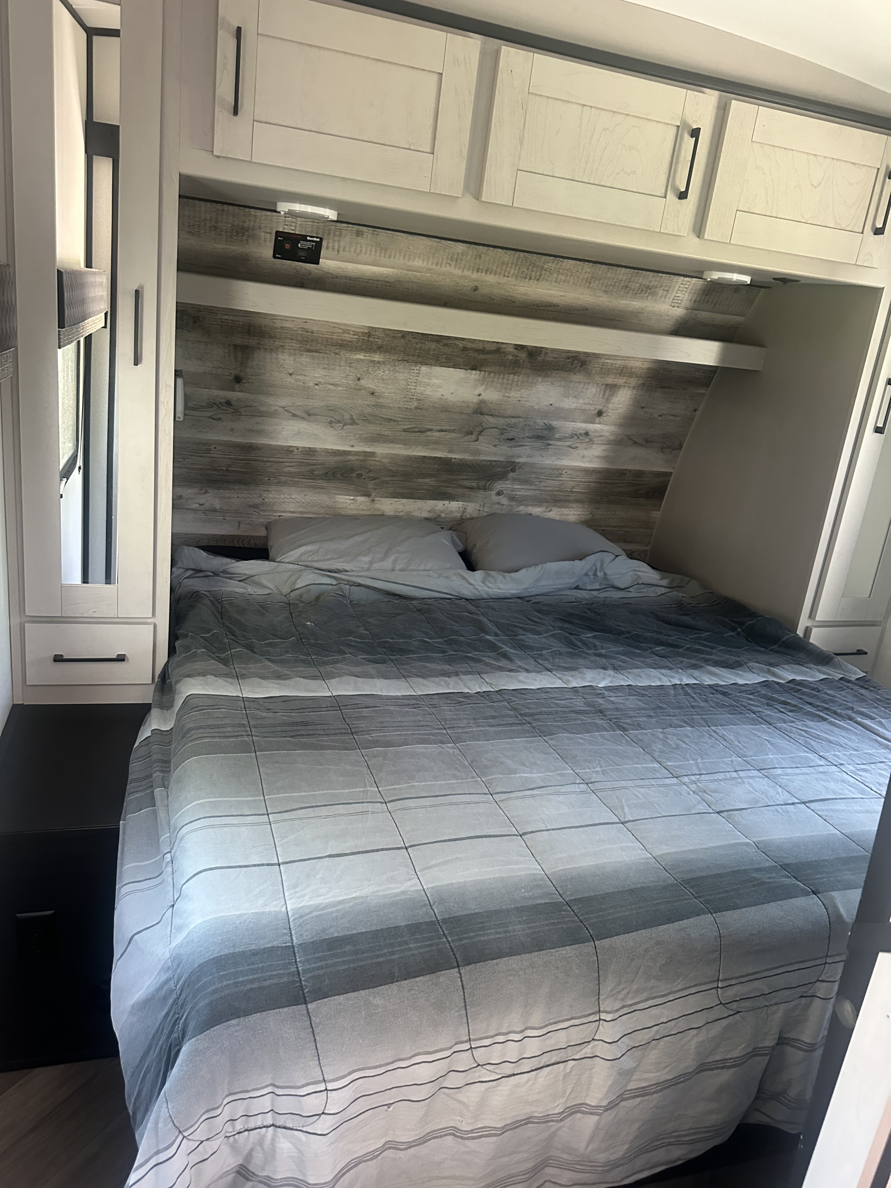 RV rental bedroom: A comfortable bed with striped comforter, wood headboard, upper cabinets, and a GenSet control panel.. Heartland RVs Torque Toy Hauler 2021