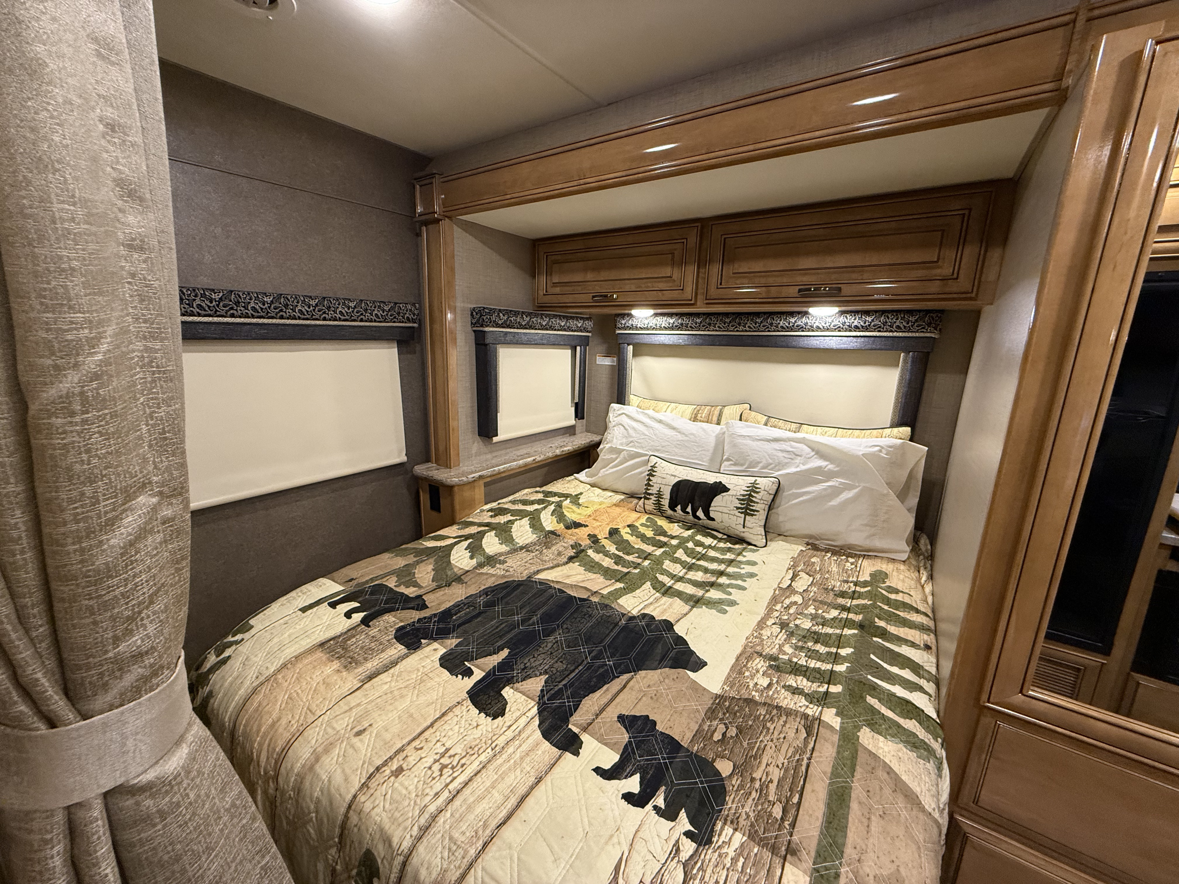 Cozy, bear-themed bedroom interior of an RV rental, featuring wooden cabinetry, headboard, and windows with blinds.. Thor Quantum 2018