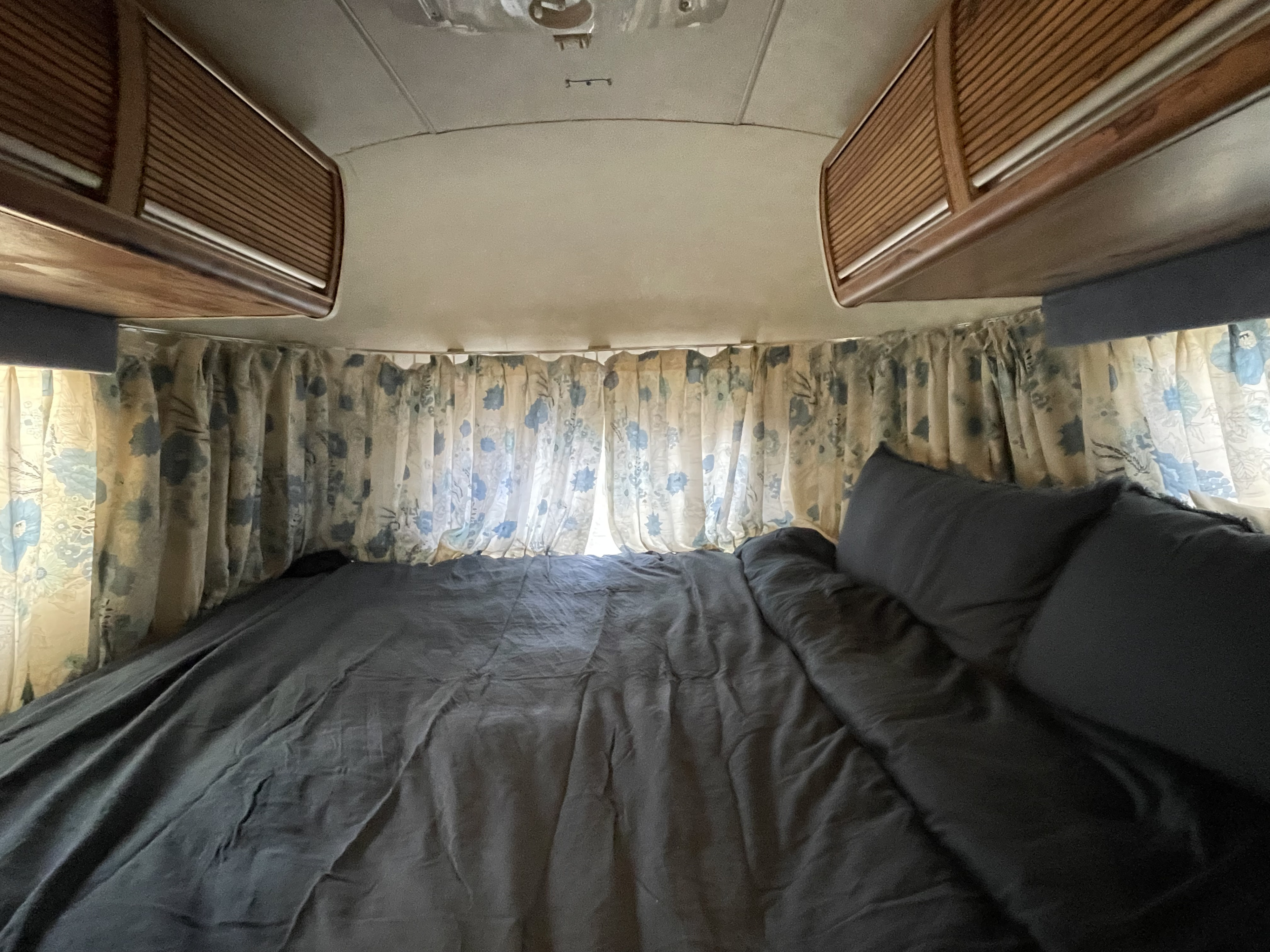 Cozy rv rental bedroom with dark bedding, floral curtains, and overhead wood cabinets, seen from the foot of the bed.. Airstream Excella 1981