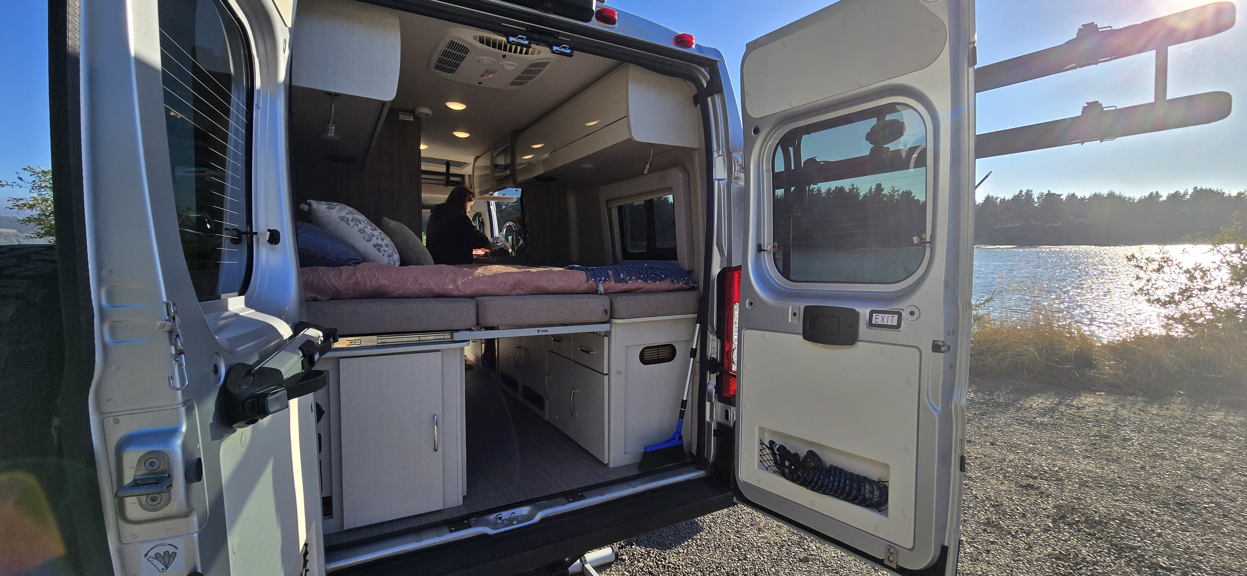 Cozy RV rental adventure awaits! Van's open rear doors reveal a comfortable bed, storage, and a person inside, with a beautiful lake view.. Thor Tellaro 2024