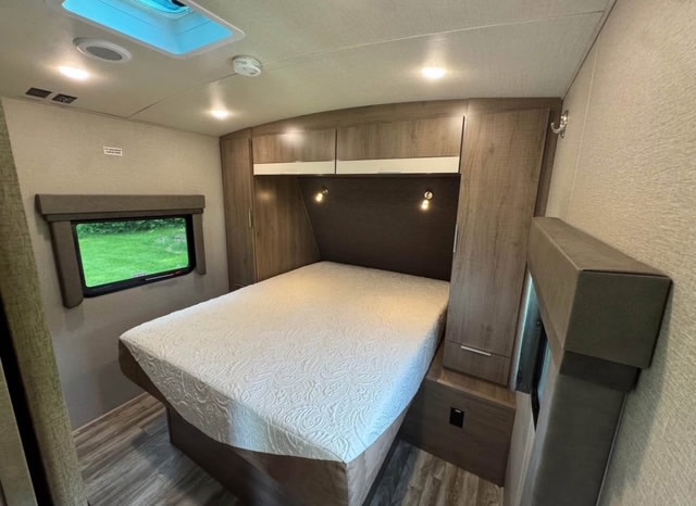 Cozy RV rental bedroom featuring a queen bed, overhead storage, and a window with a green outdoor view.. Grand Design Imagine 2020