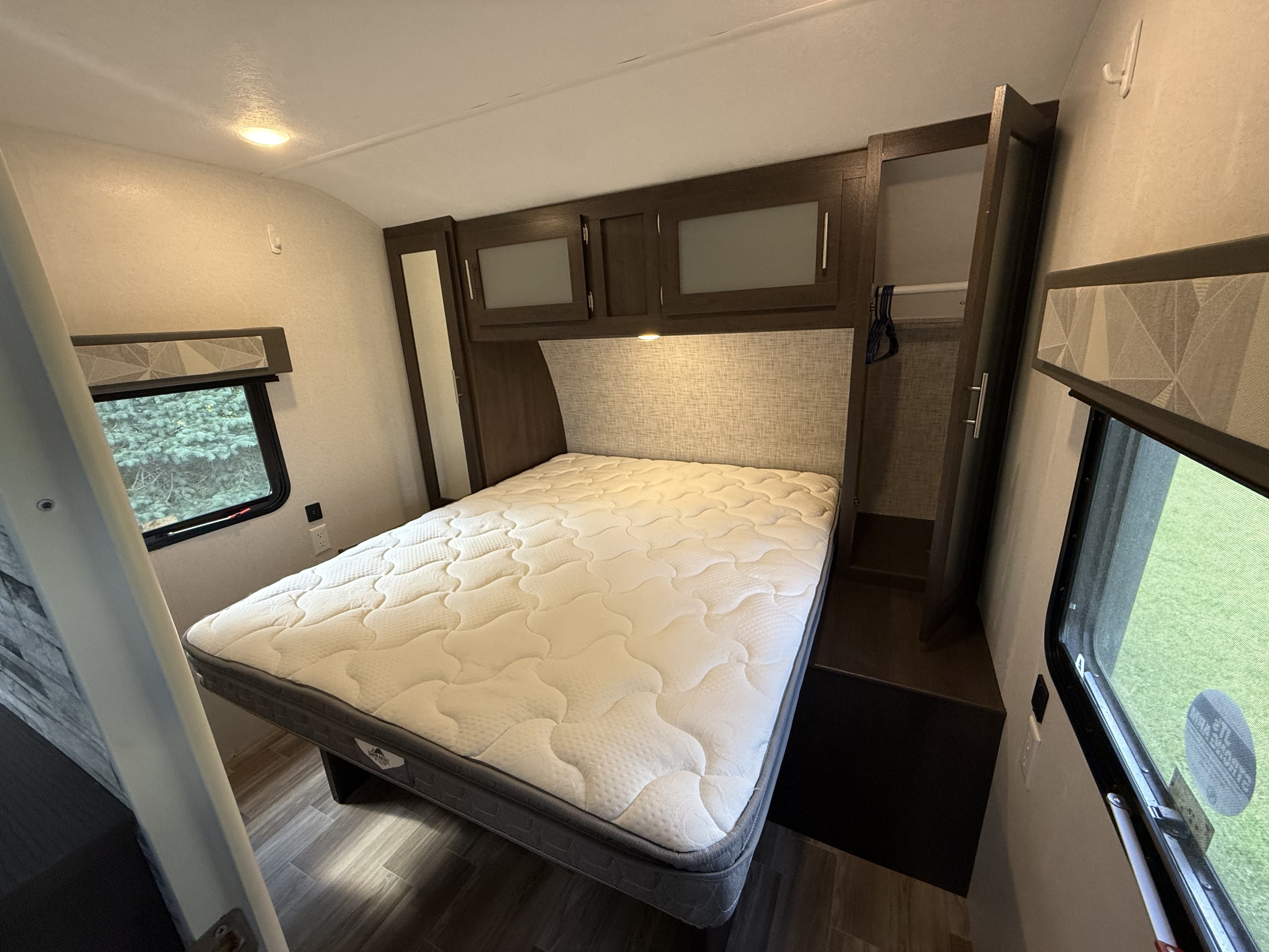 A comfortable, clean RV rental bedroom featuring a white mattress, dark wood overhead cabinets, and a window overlooking greenery.. Forest River Salem 2019