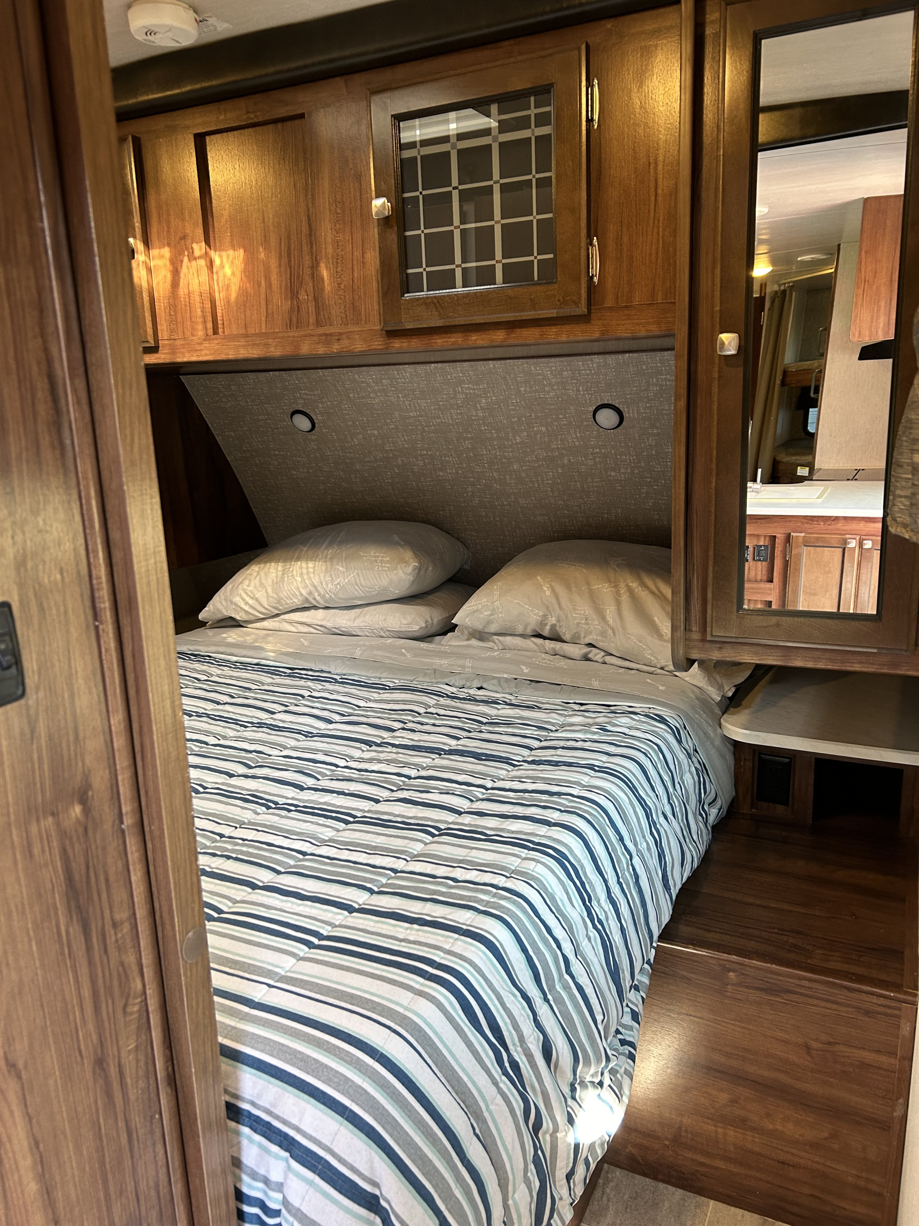 This RV rental bedroom features a striped bed, lighted headboard, and wood cabinetry with a mirrored door.. Heartland RVs Mallard 2019