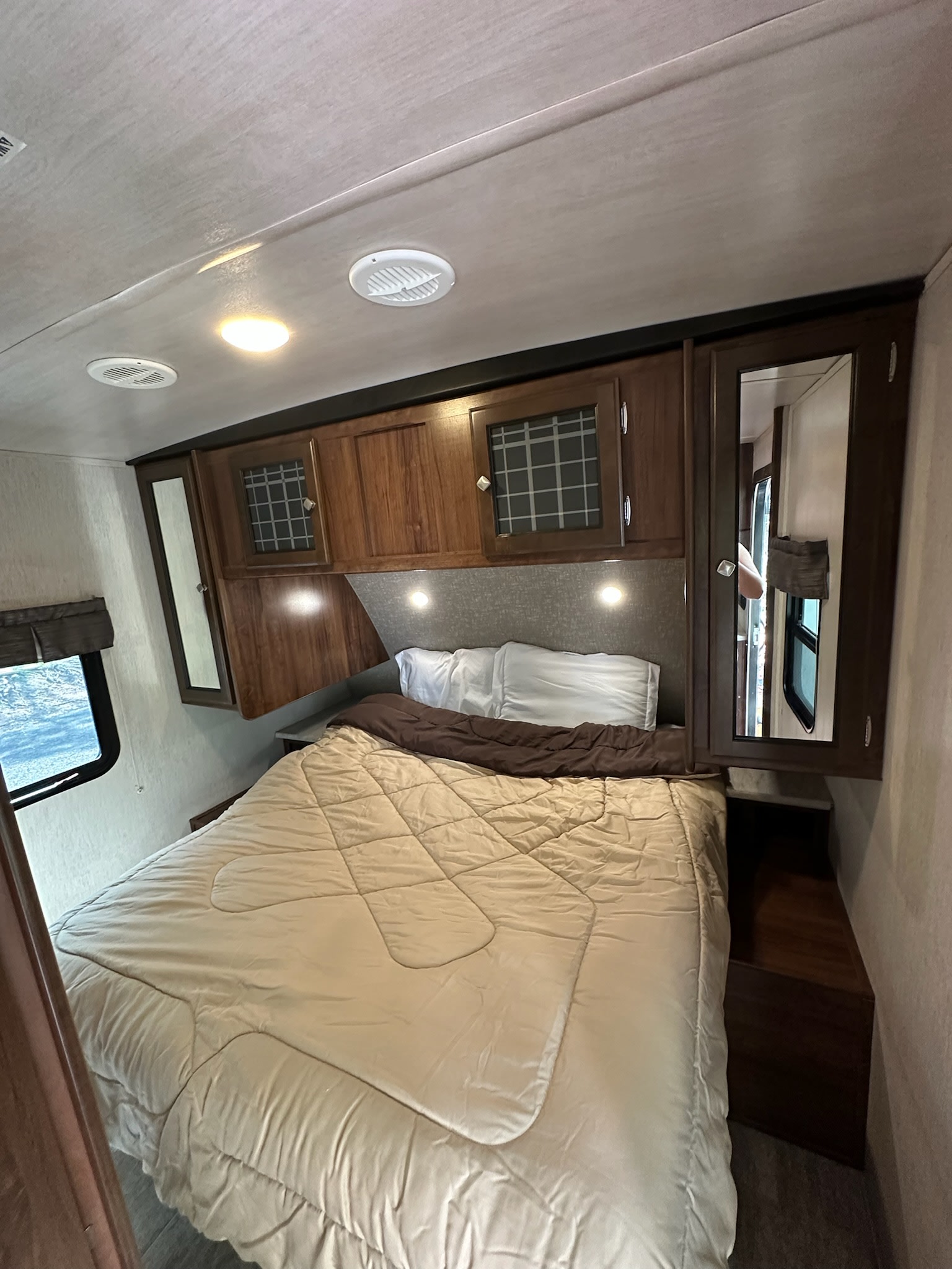 This cozy RV rental bedroom features a comfy bed, dark wood overhead cabinets, and convenient lighting, perfect for travel.. Heartland RVs Mallard 2019