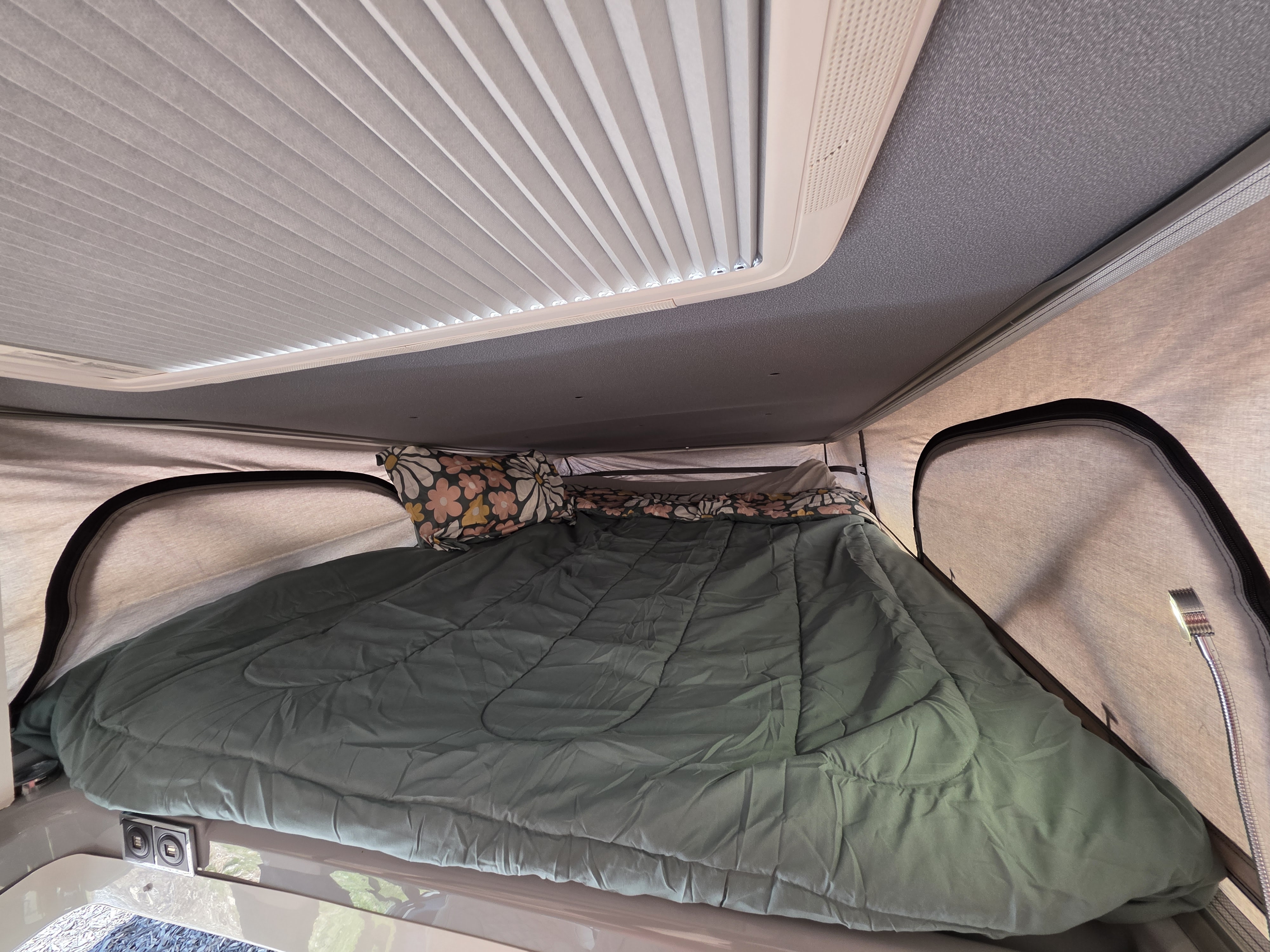 Inside an RV rental, a comfy pop-top bed with green bedding, floral pillow, and integrated charging ports.. Jayco Swift 2024