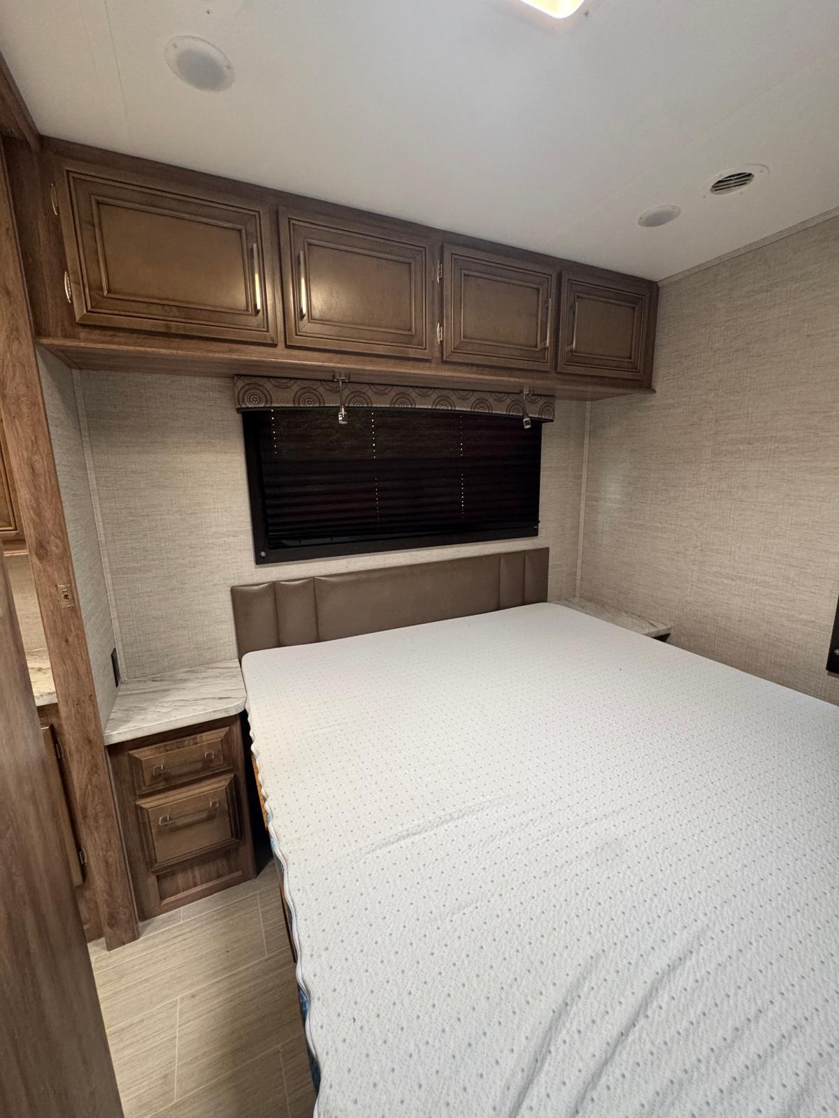 This RV rental bedroom features a large bed, dark wood overhead cabinets, and built-in nightstands with window blinds.. Entegra Coach Odyssey 2021