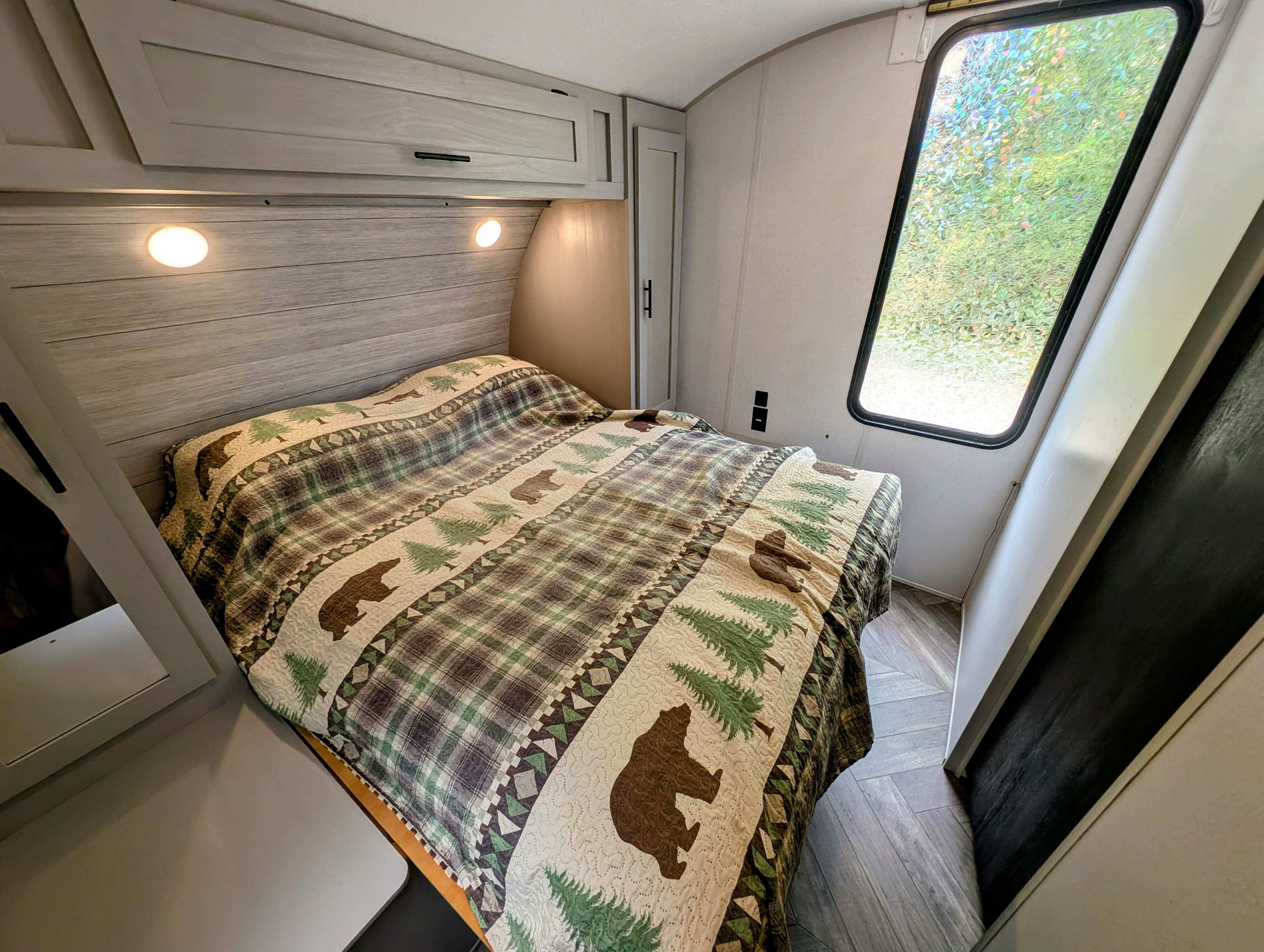 Cozy RV rental bedroom featuring a bear and tree-themed quilt, overhead cabinets, reading lights, and a decorative window.. Forest River Salem 2021