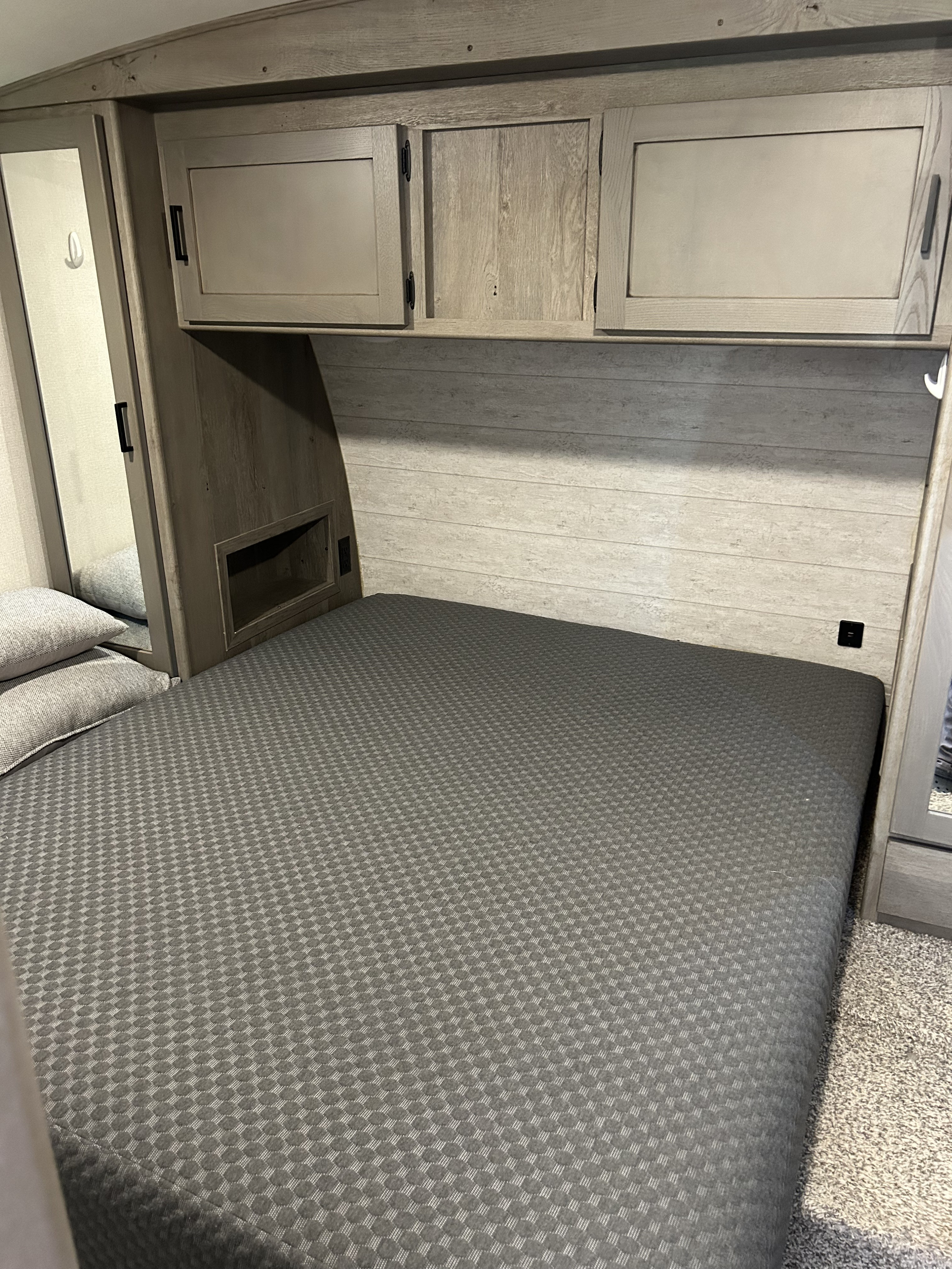 Compact RV rental bedroom featuring a grey bed, overhead cabinets, a mirrored wardrobe, and cozy wood-paneled walls.. Keystone Outback 2021