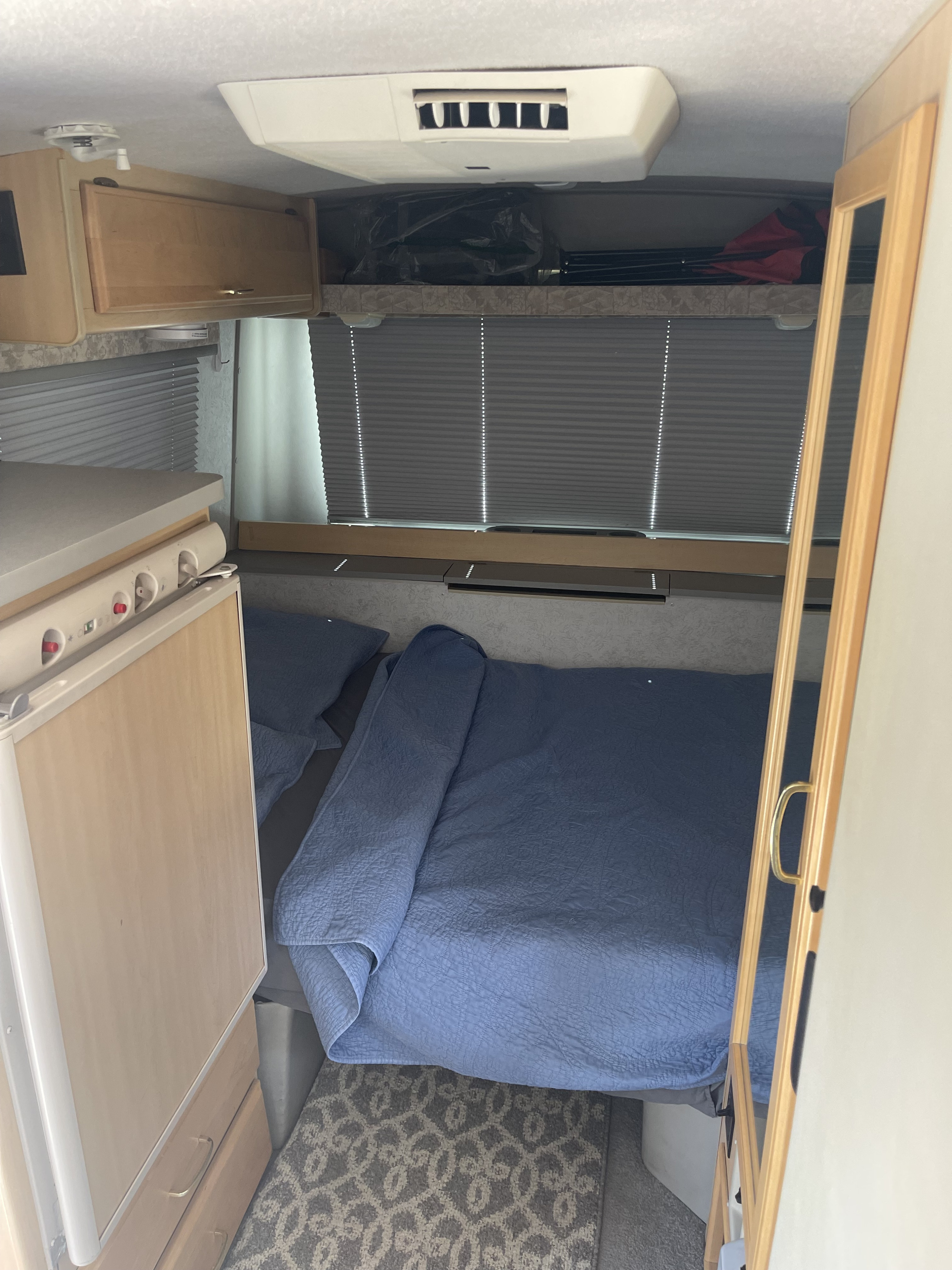 Cozy RV rental interior showing a bed with blue bedding, a small kitchen, and overhead storage. Ideal for travel.. Eurovan Camper Rialta 2000