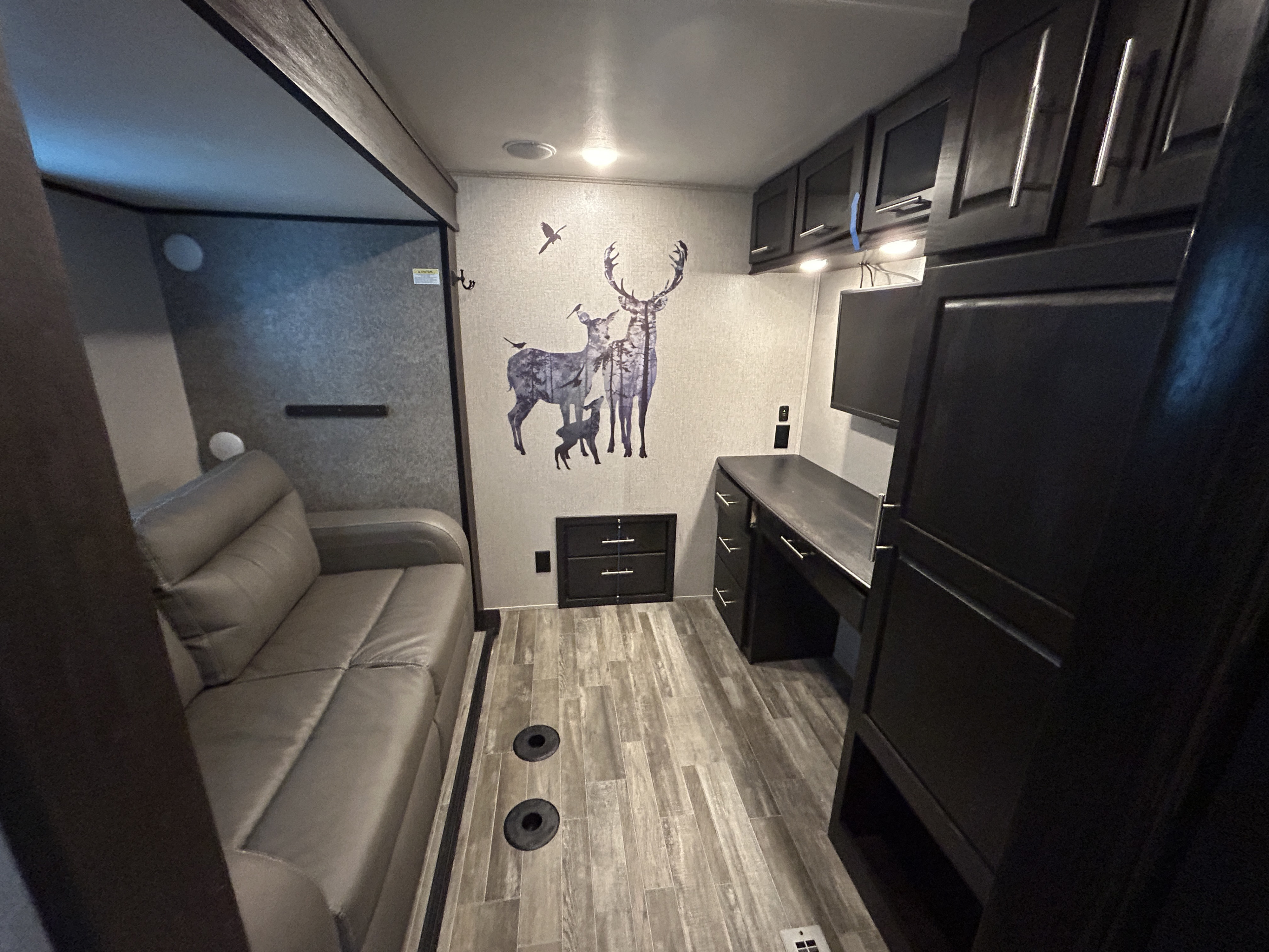 This RV rental features a versatile living space with a grey sofa, desk, overhead cabinets, wood-look floor, and deer wall art.. Jayco North Point 2021
