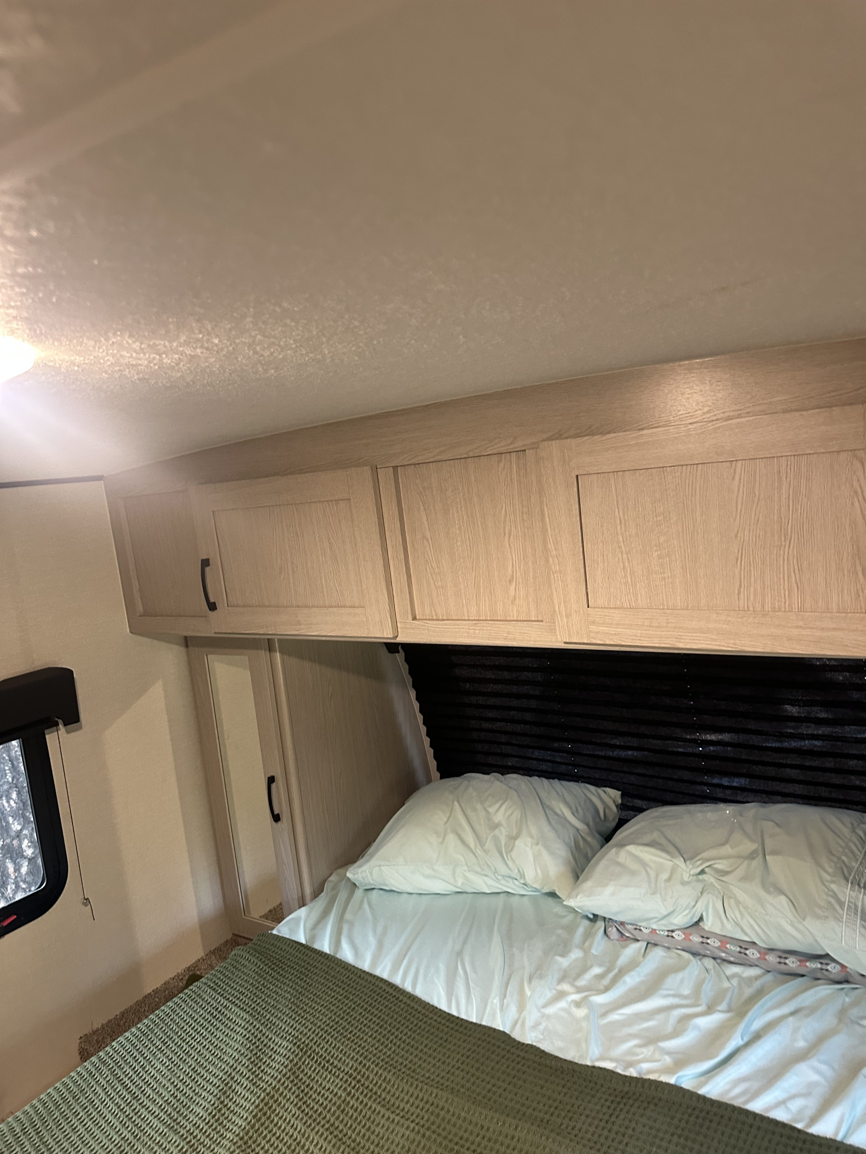 RV rental bedroom featuring a cozy bed with light blue sheets, overhead light wood cabinets, and a window.. Coachmen Apex Nano - Off Grid 2022