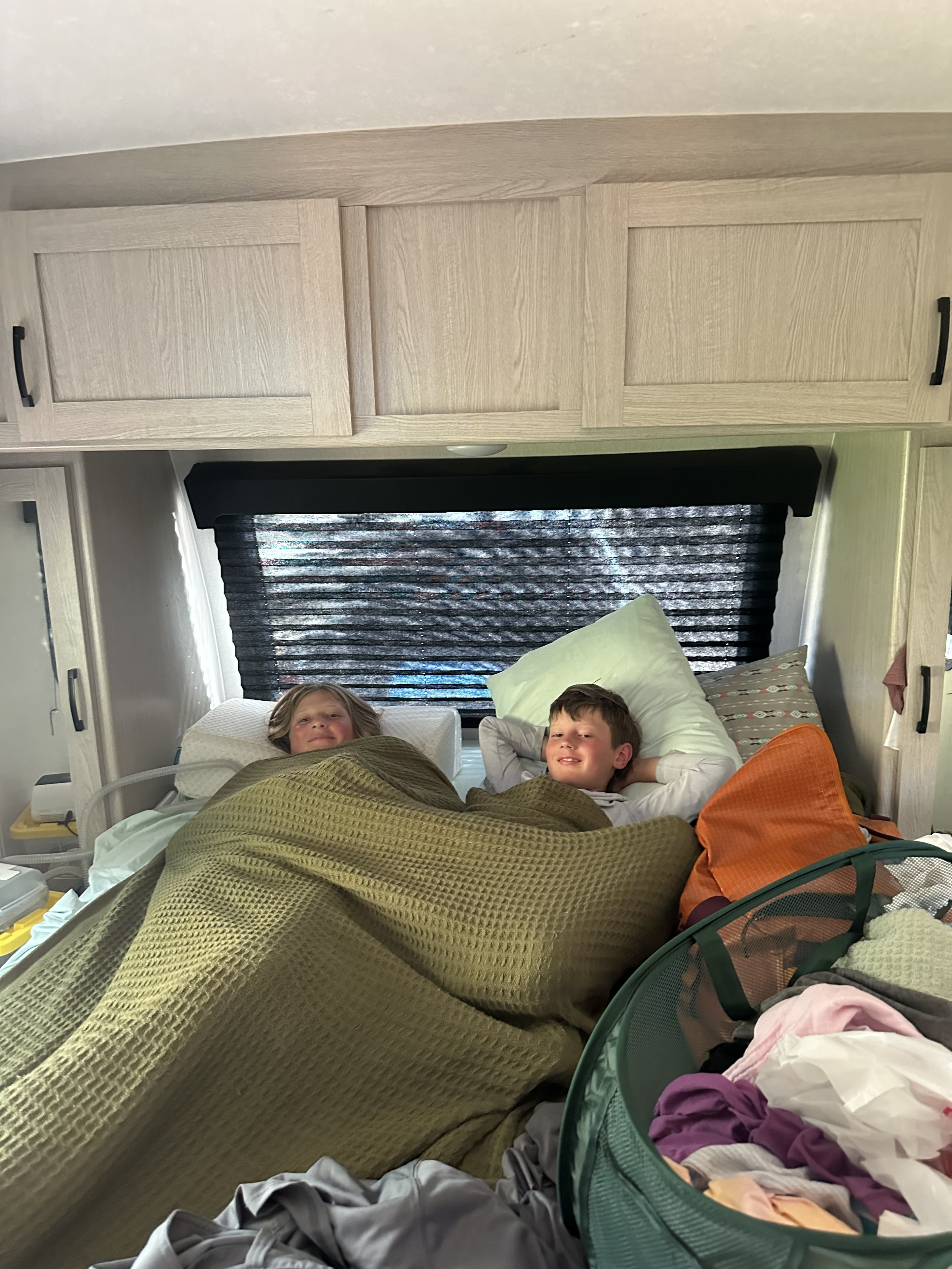 Smiling boys relax comfortably in their RV rental bunk, covered by a green blanket. Perfect for family travel and cozy adventures.. Coachmen Apex Nano - Off Grid 2022