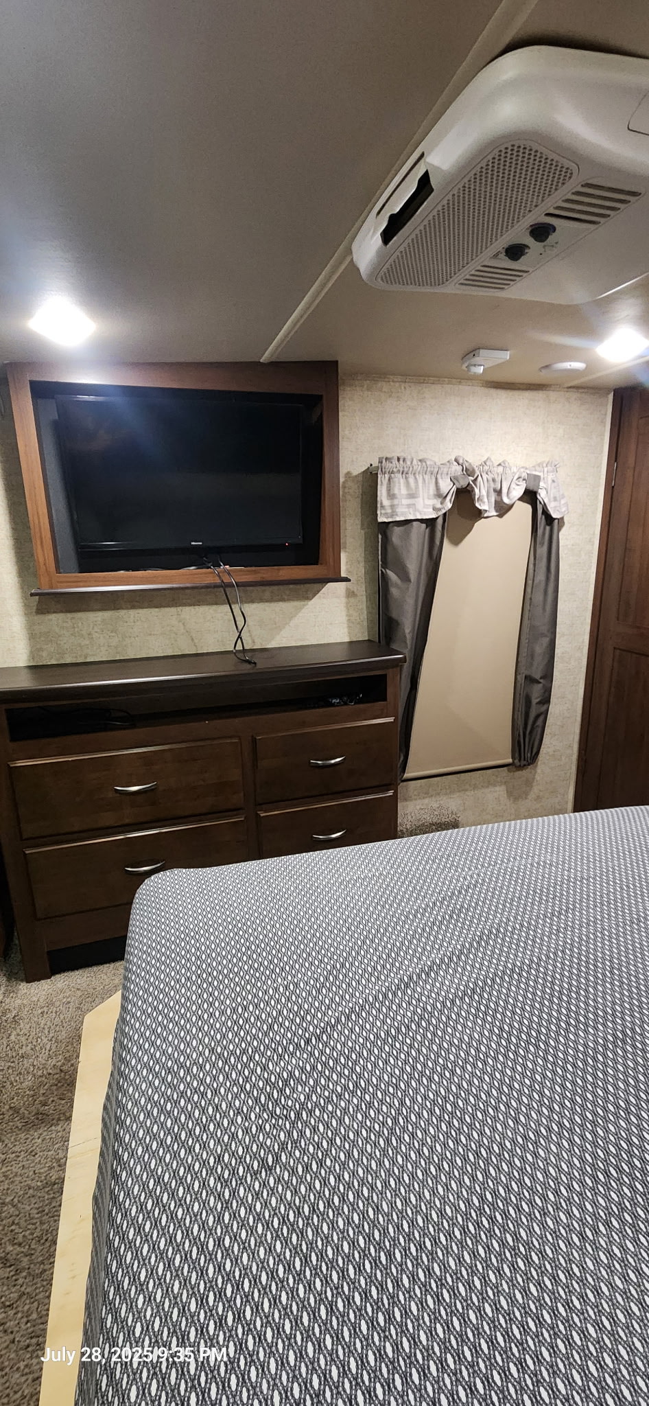 RV rental bedroom with a mounted TV, wooden dresser, bed with patterned cover, and ceiling AC unit.. Forest River Sandpiper 2017