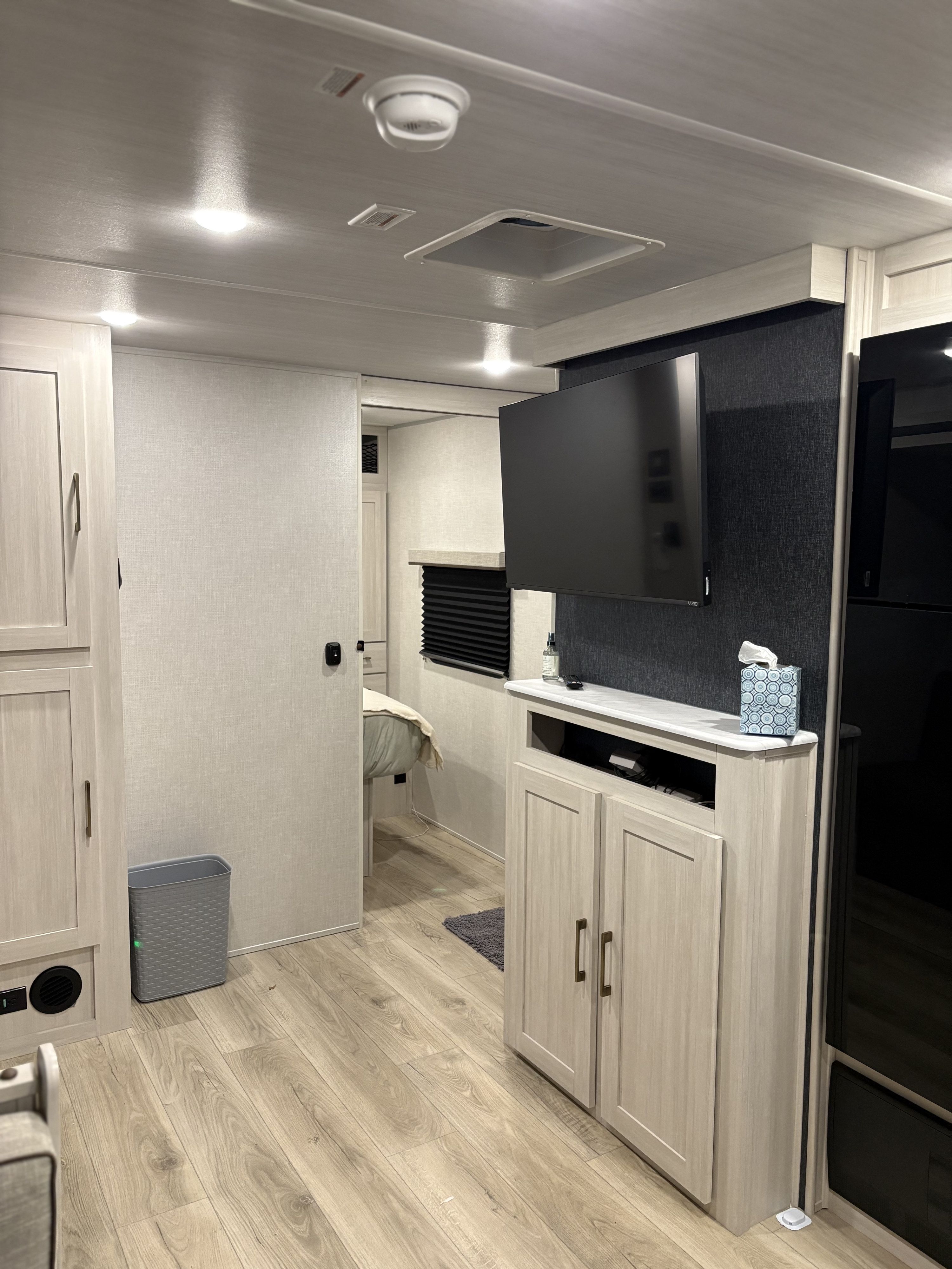 Modern RV rental interior showing a light wood living area with TV, cabinets, and a sliding door leading to a clean bedroom.. Surveyor Legend X 27CAMP 2025