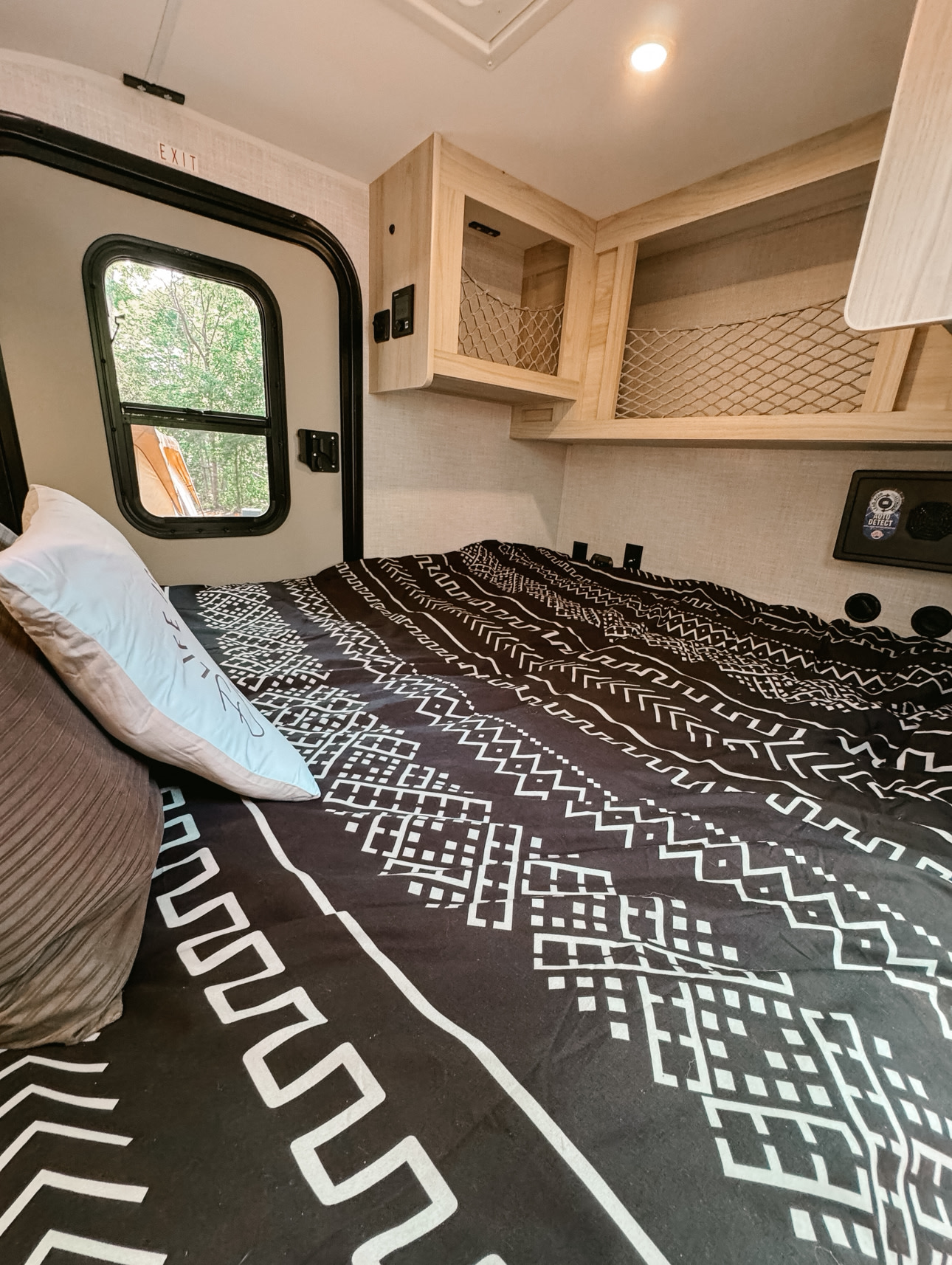 Cozy RV rental interior featuring a bed with black patterned bedding, wooden overhead storage, and an "EXIT" sign by the door.. Skinny Guy Campers Armor 10LT 2025