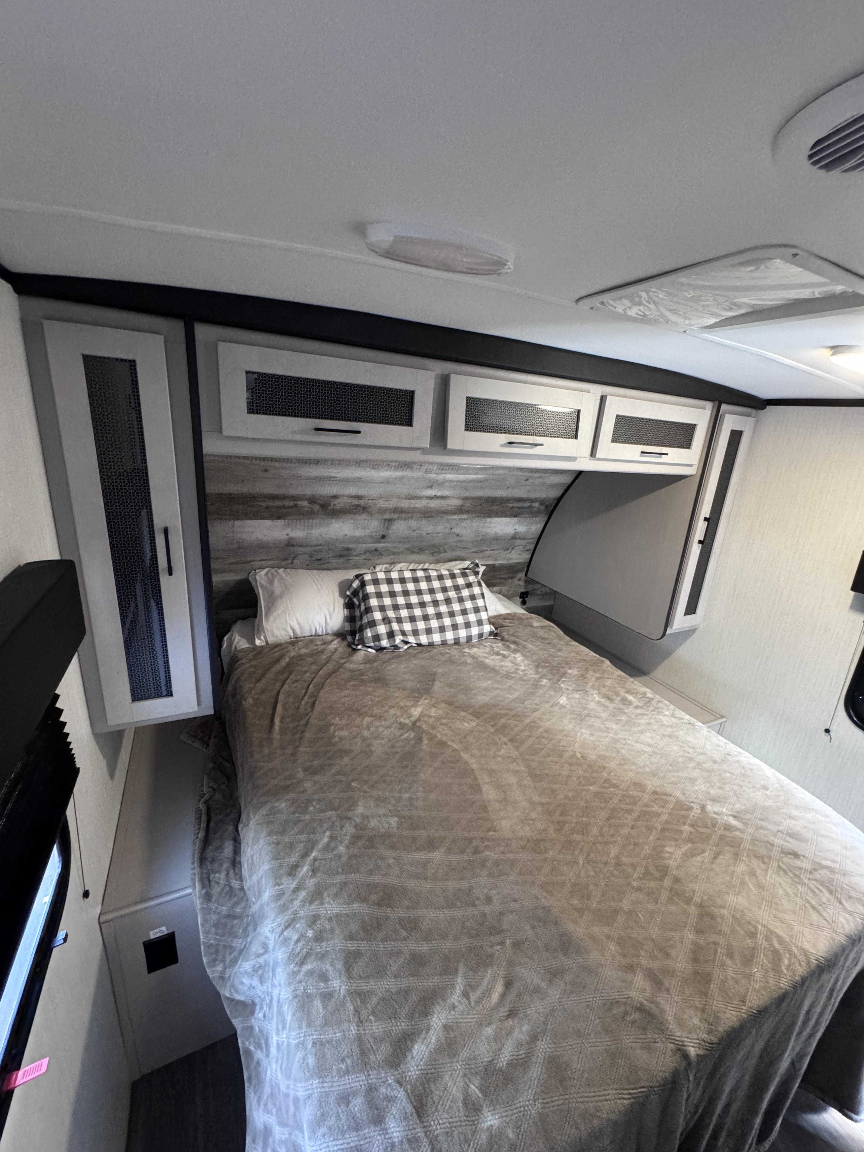 Cozy RV rental bedroom with a bed, gray quilted blanket, wood-look headboard, and light-colored overhead cabinets.. Heartland RVs Mallard 2024