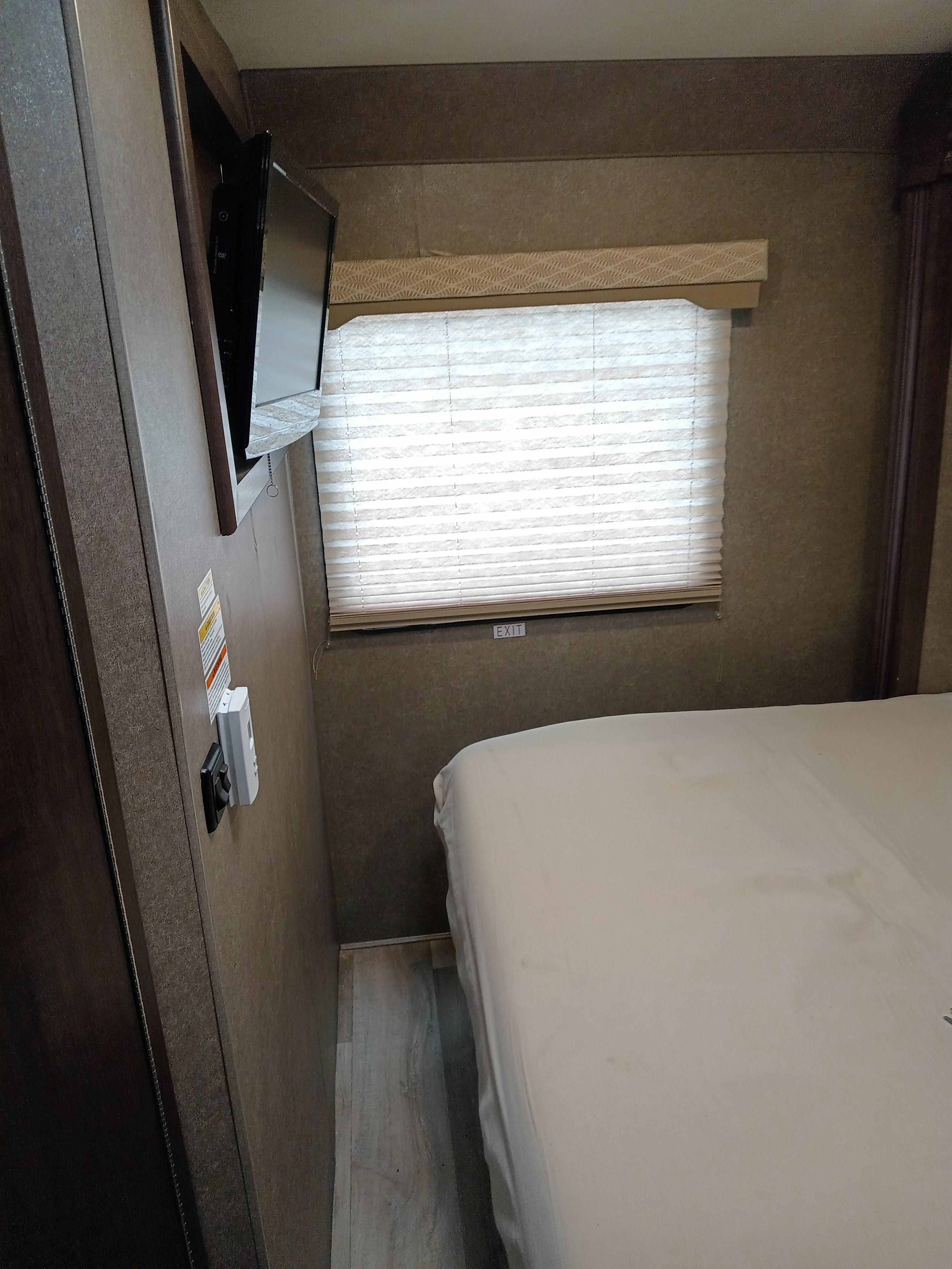 RV rental bedroom: White bed, wall-mounted TV, window with blinds. An 'EXIT' sign is visible below the window.. Thor Freedom Elite 2020