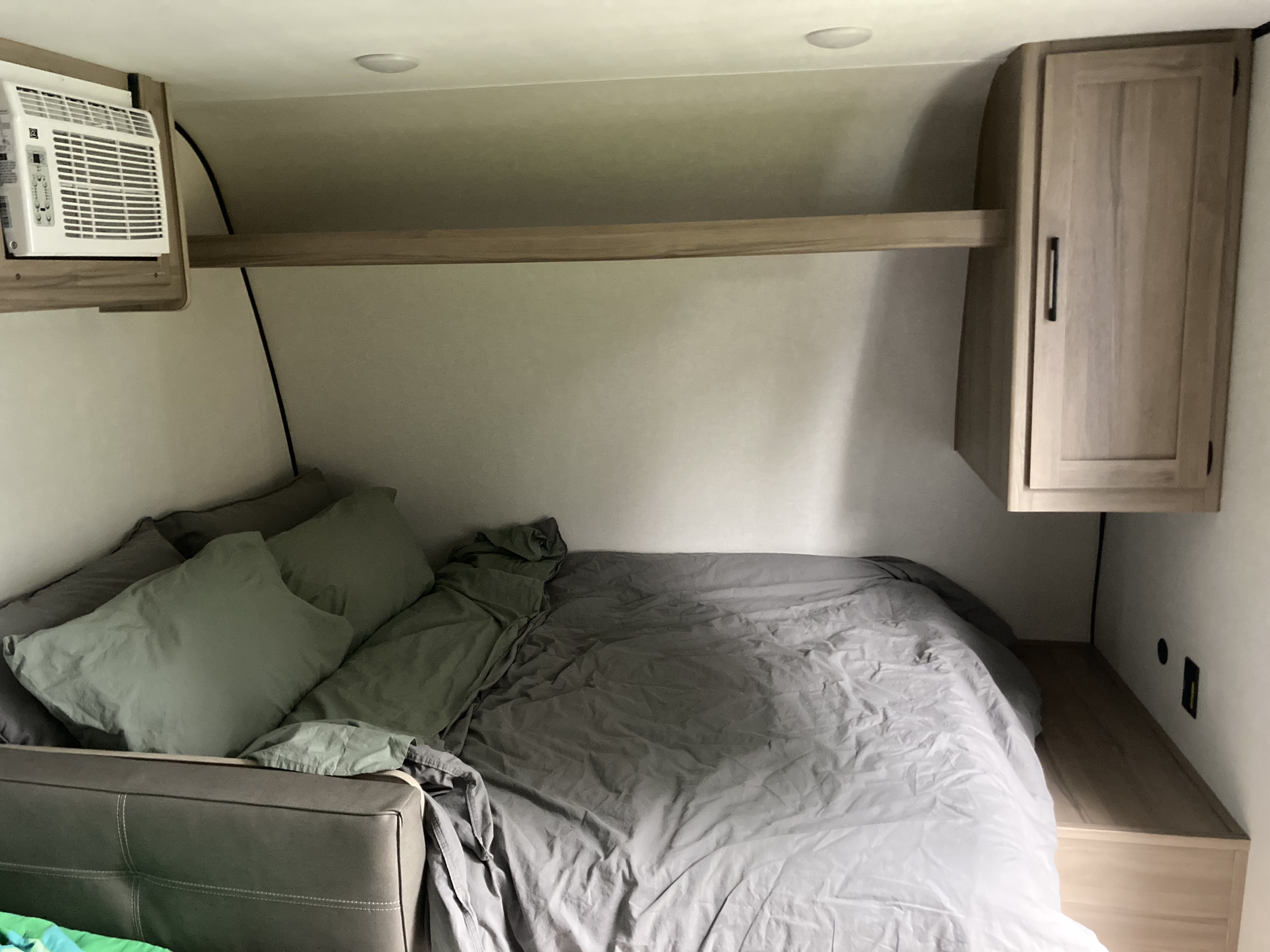 Cozy RV rental bedroom featuring a full bed with light bedding, a mounted AC unit, and an overhead wooden cabinet. Ideal for travel.. Keystone Coleman 17b 2025
