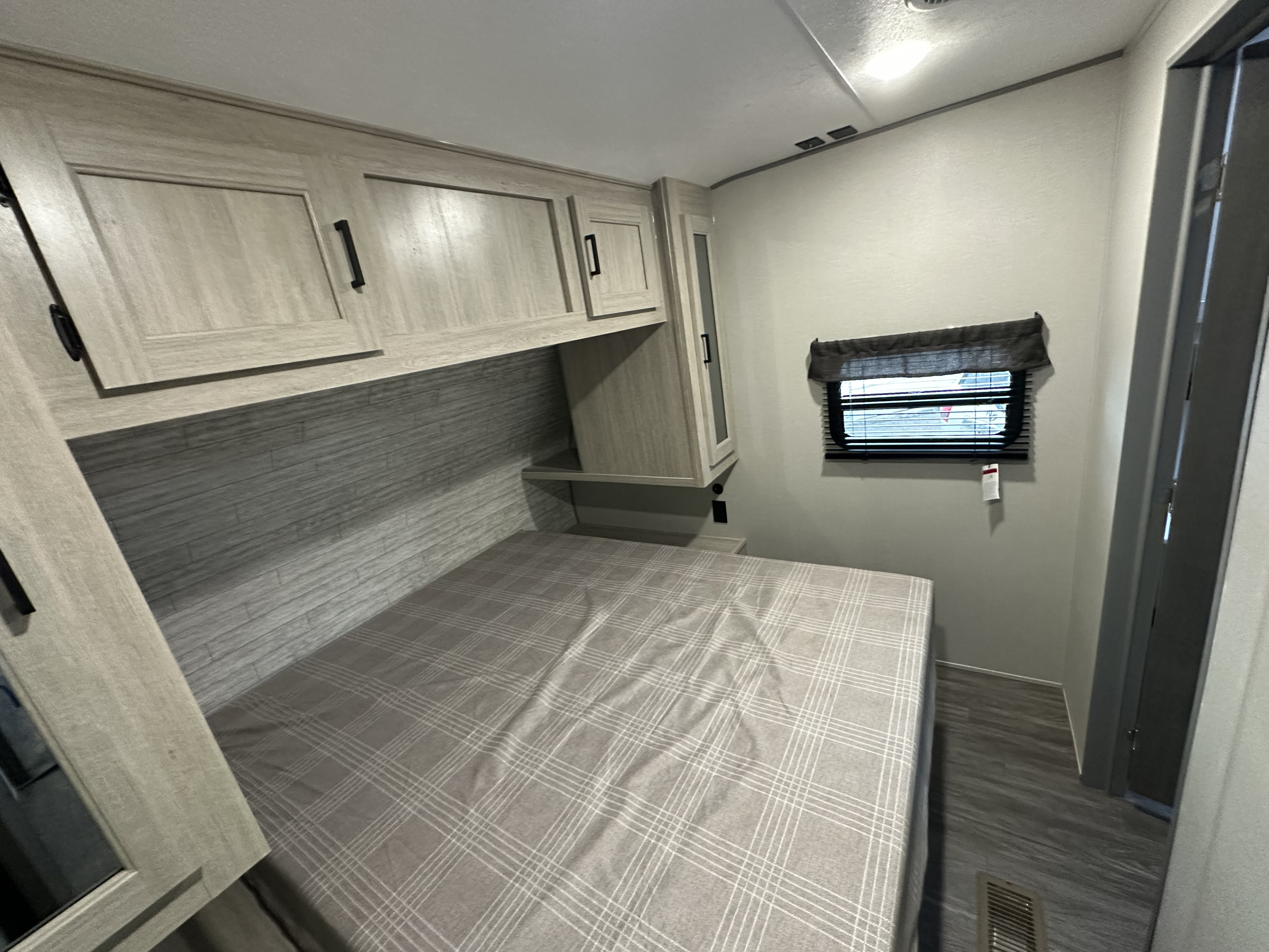 RV rental bedroom with plaid bed, overhead cabinets, window with blinds, and wood-look flooring.. Forest River Aurora 29TQS 2026