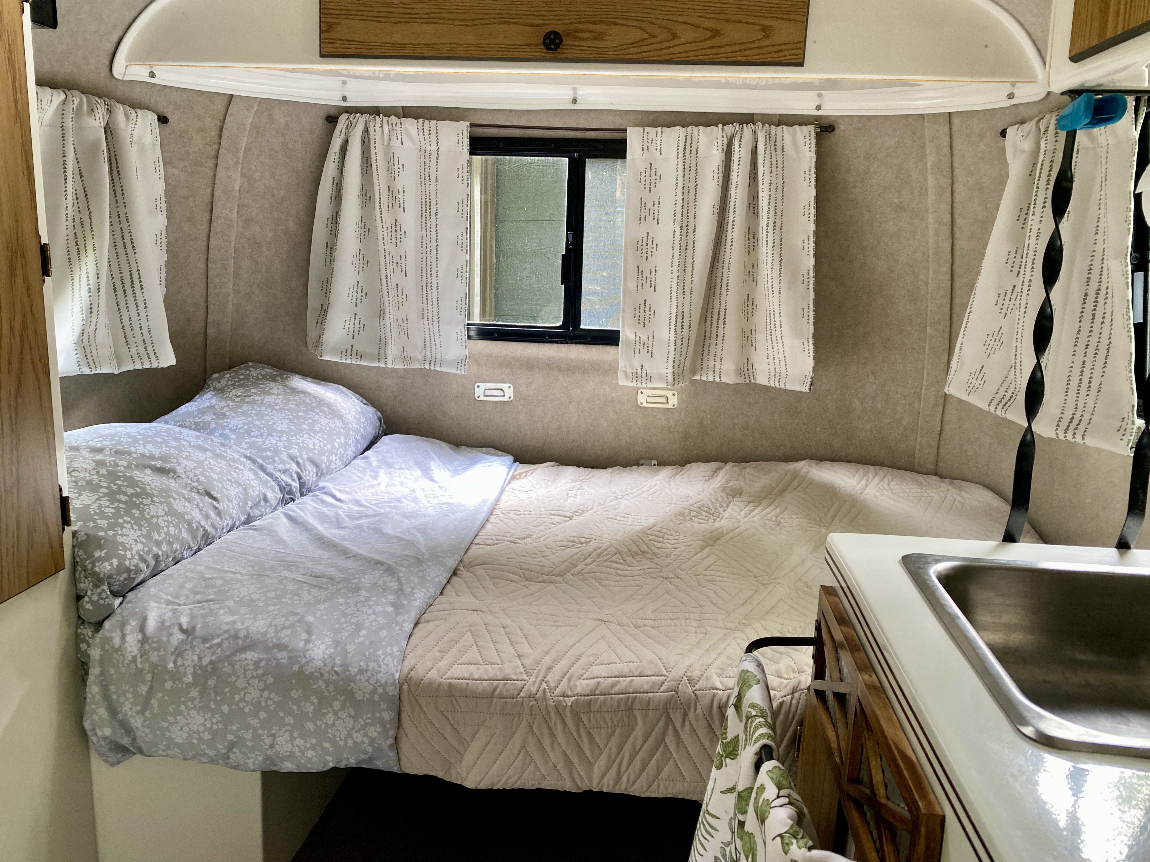 Cozy rv rental interior: comfortable bed with floral and quilted bedding, windows with curtains, and a compact sink.. Scamp Scamp Trailer 1999