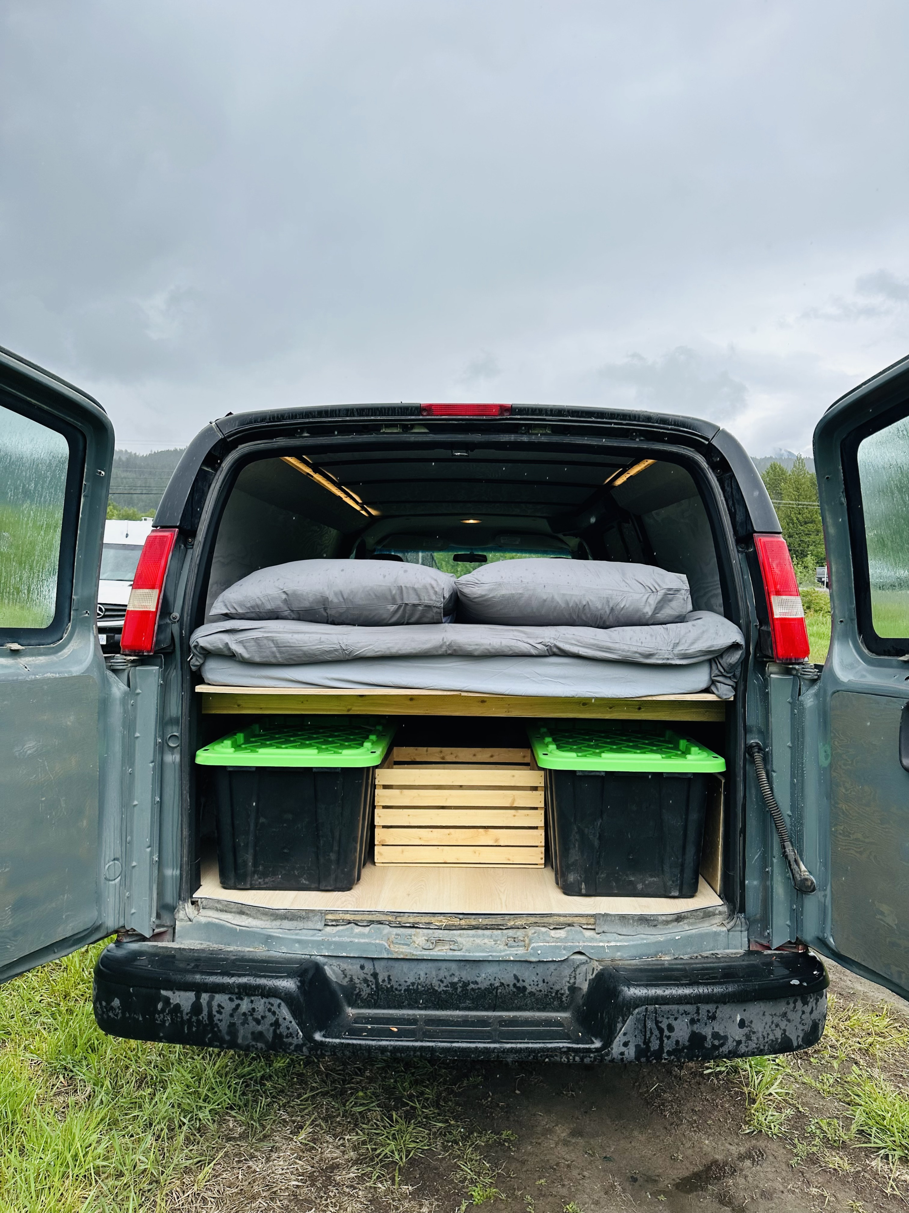 Rear view of a converted rv rental van with open doors, showing a raised bed, pillows, and under-bed storage for camping.. Chevrolet Express 2003