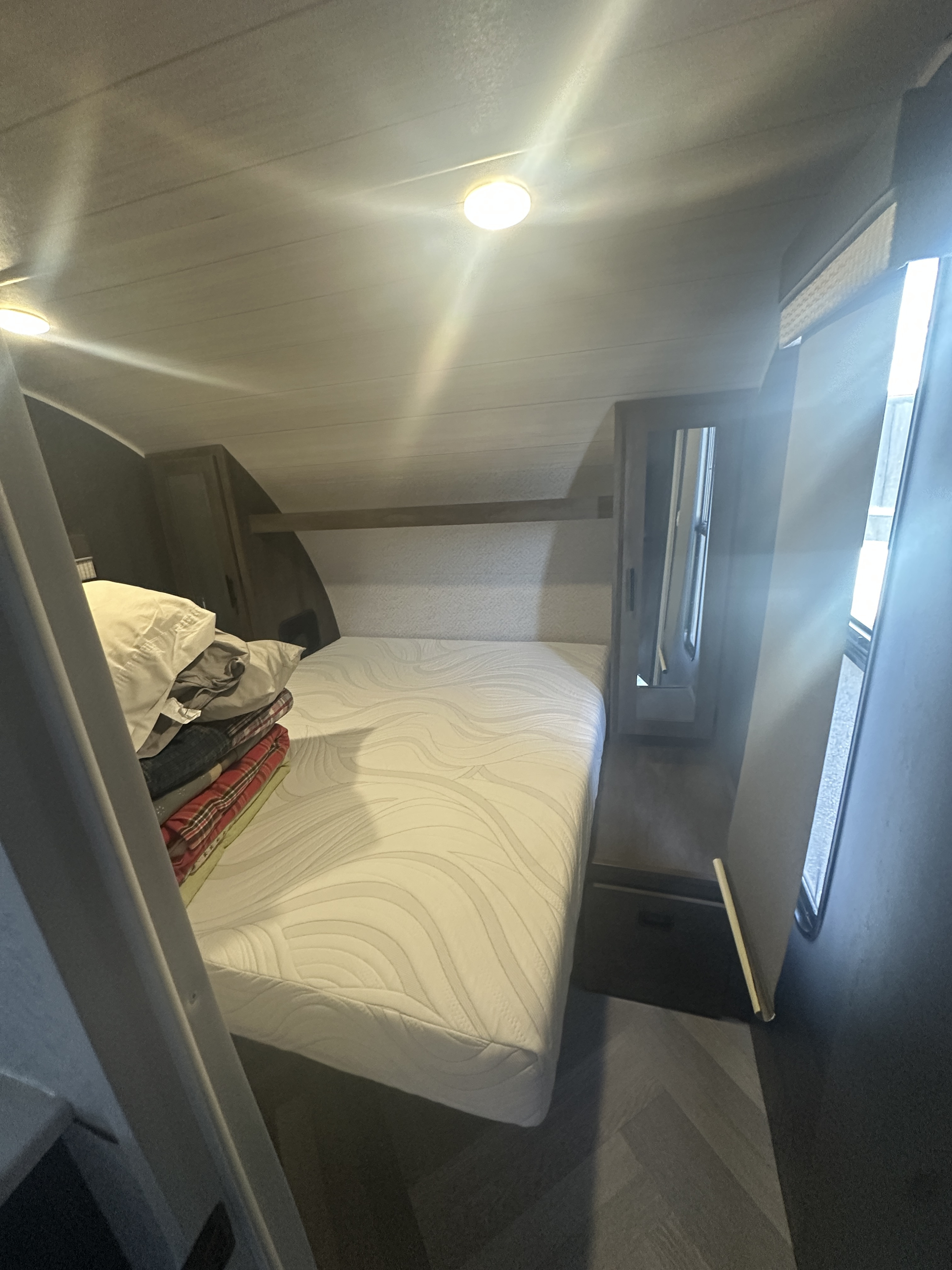 Inside an RV rental: a compact bedroom with a white mattress, stacked linens, overhead storage, and a bright window.. Forest River Salem Cruise Lite 2022